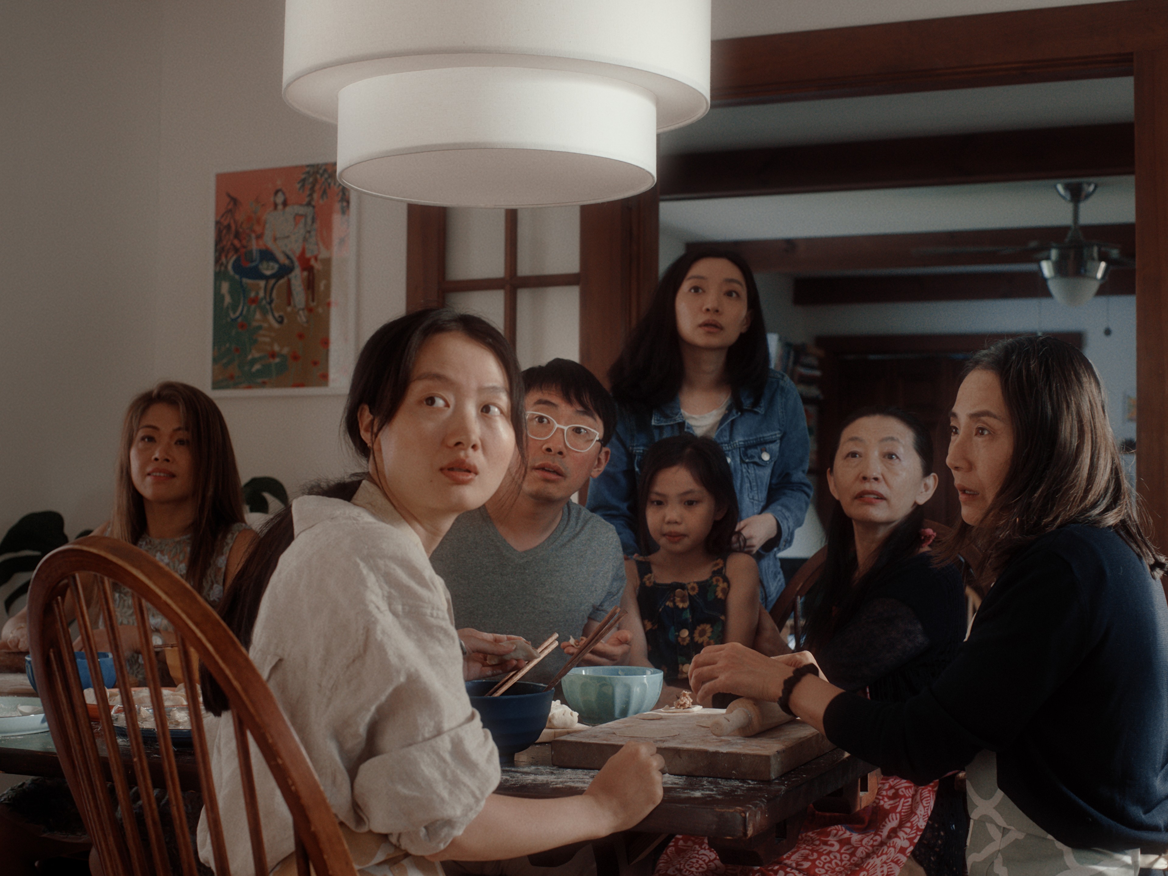 A group of adults and children gathered around a dining table, all looking toward something off-camera.