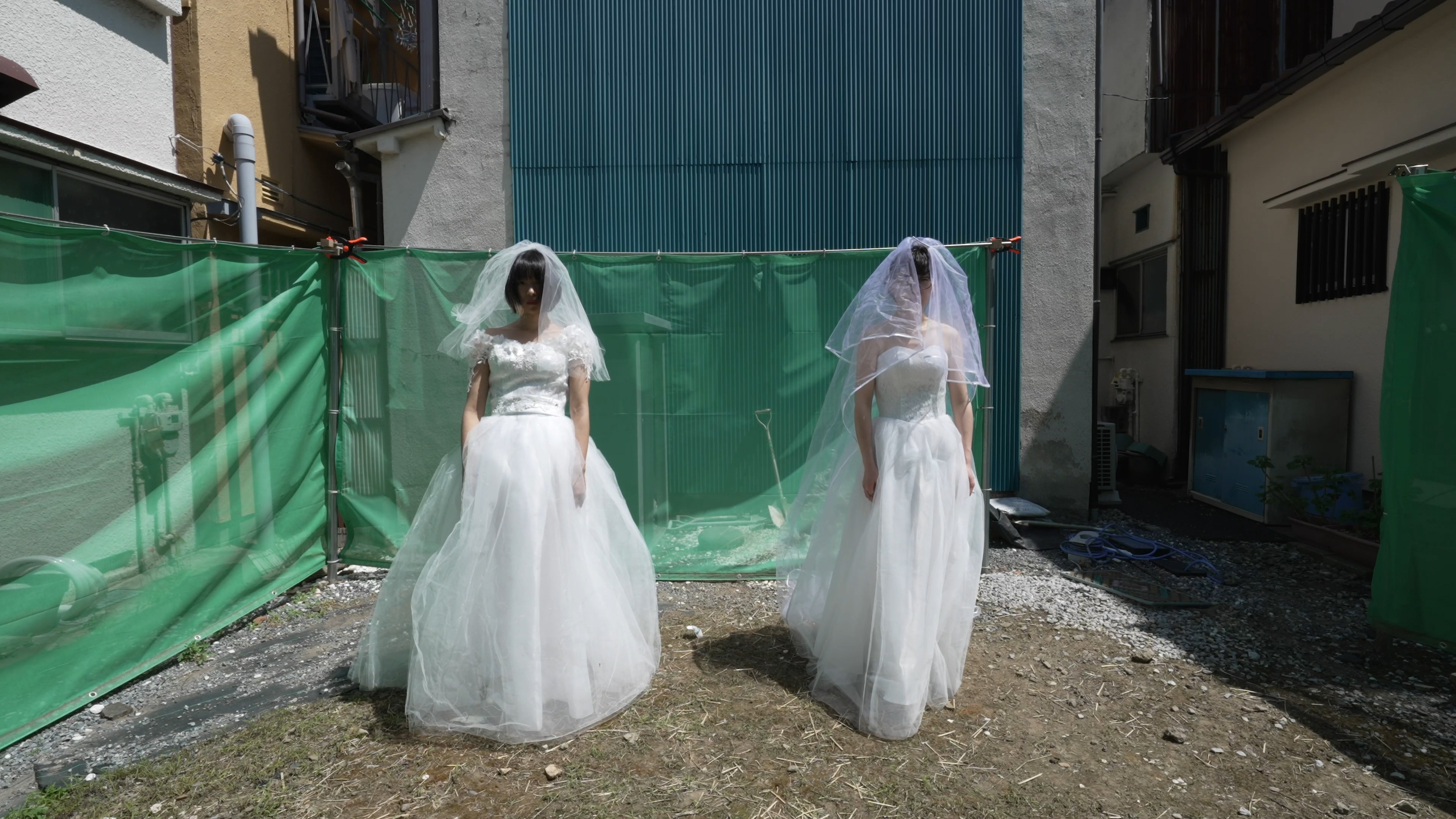 Two figures in white wedding dresses and veils stand in a sunlit courtyard enclosed by green construction fencing and industrial buildings.