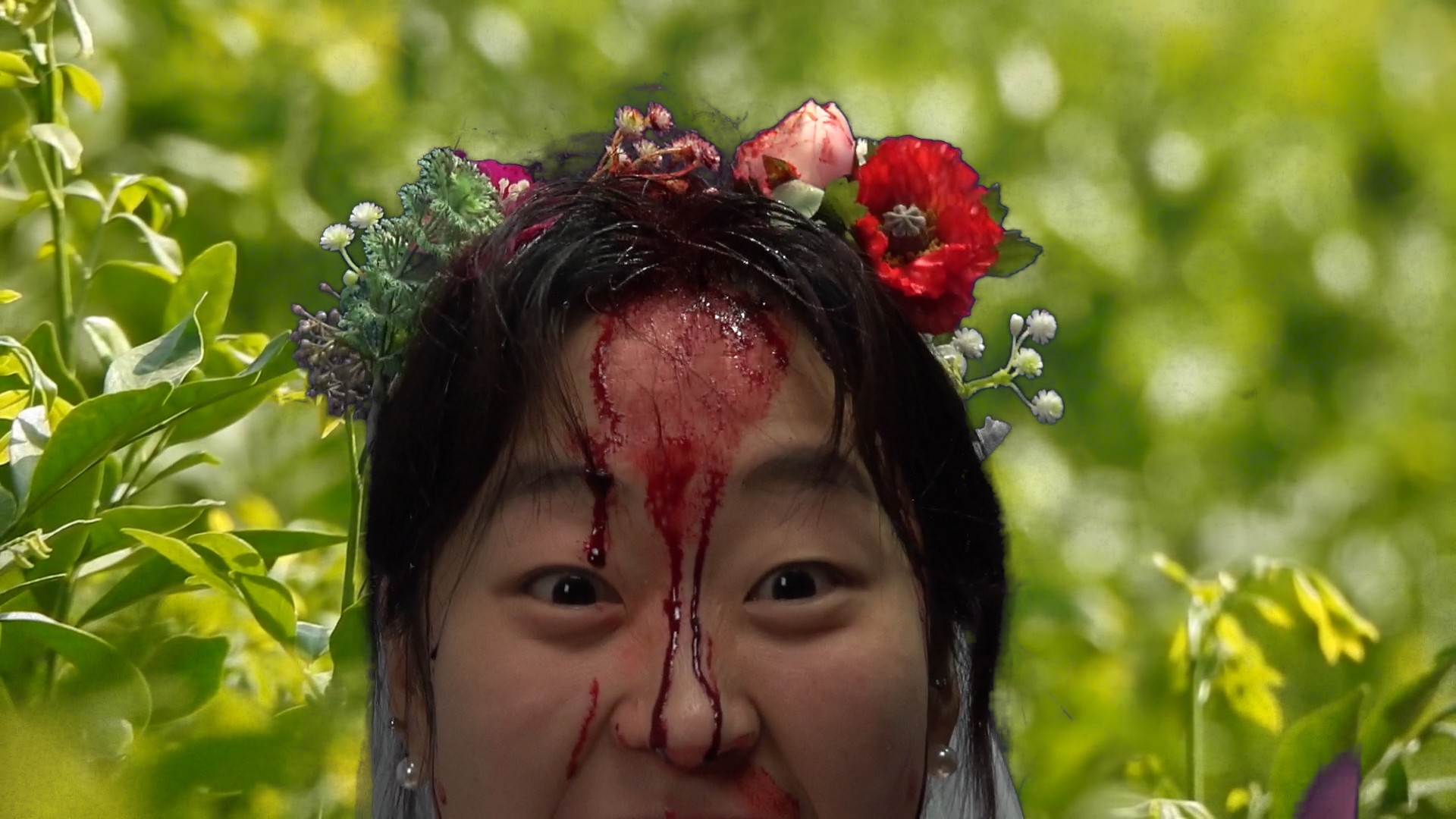 A woman wearing a floral crown looks directly at the camera, with blood running down her forehead and face against a backdrop of green foliage.