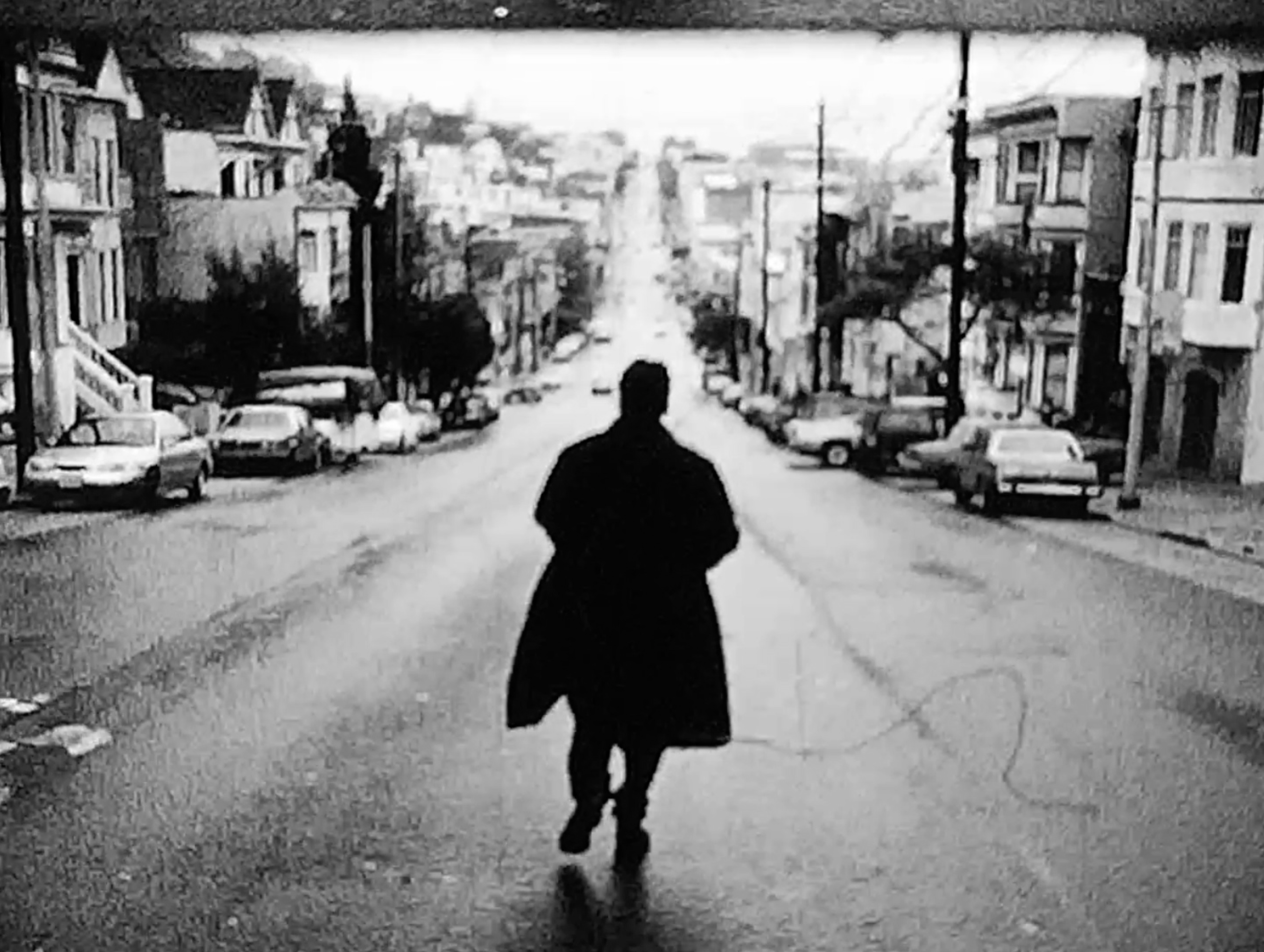 A solitary figure walking down the middle of a steep city street in a grainy black-and-white image.