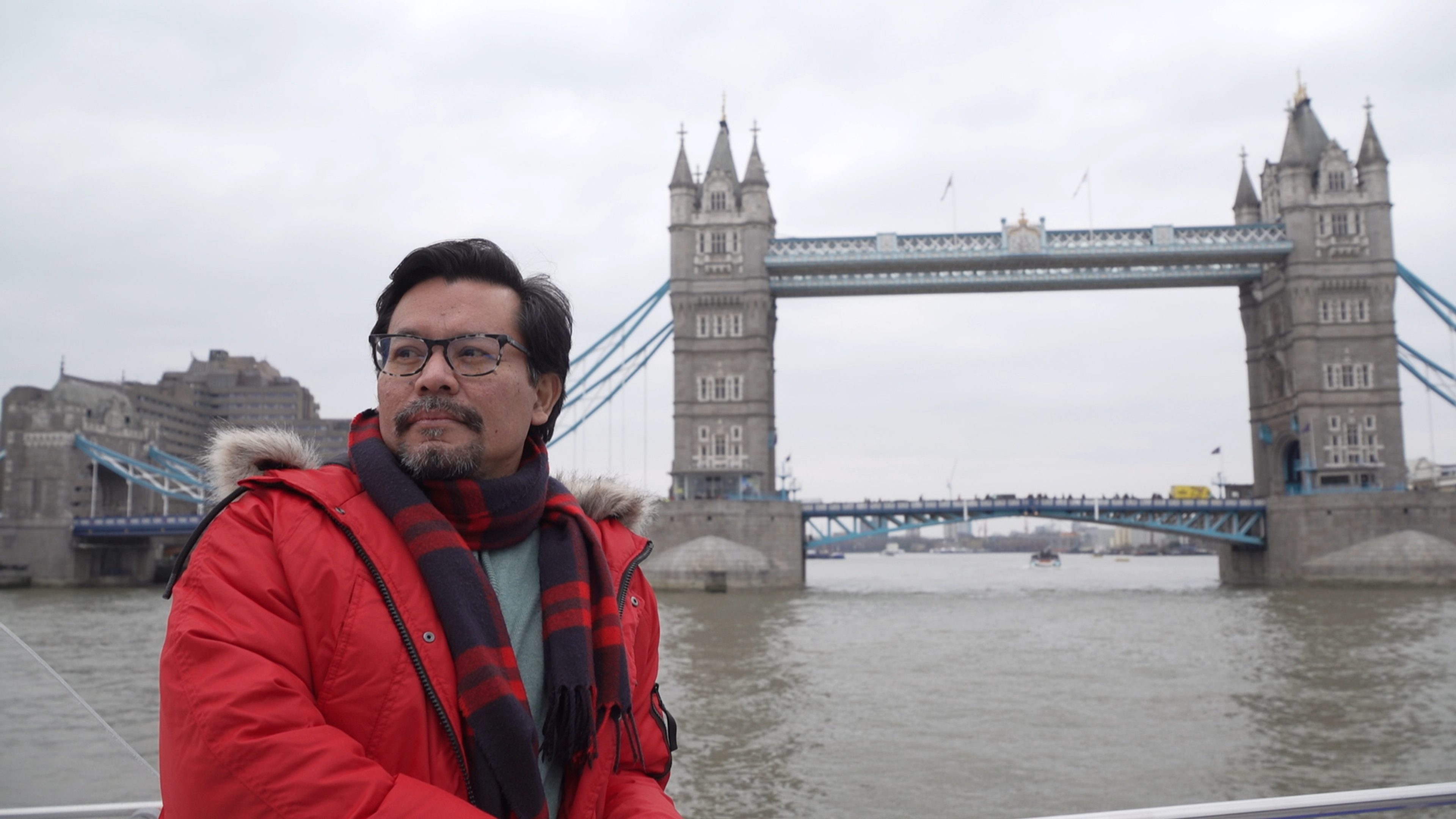 A person wearing glasses and a red coat and scarf next to Tower Bridge in London.