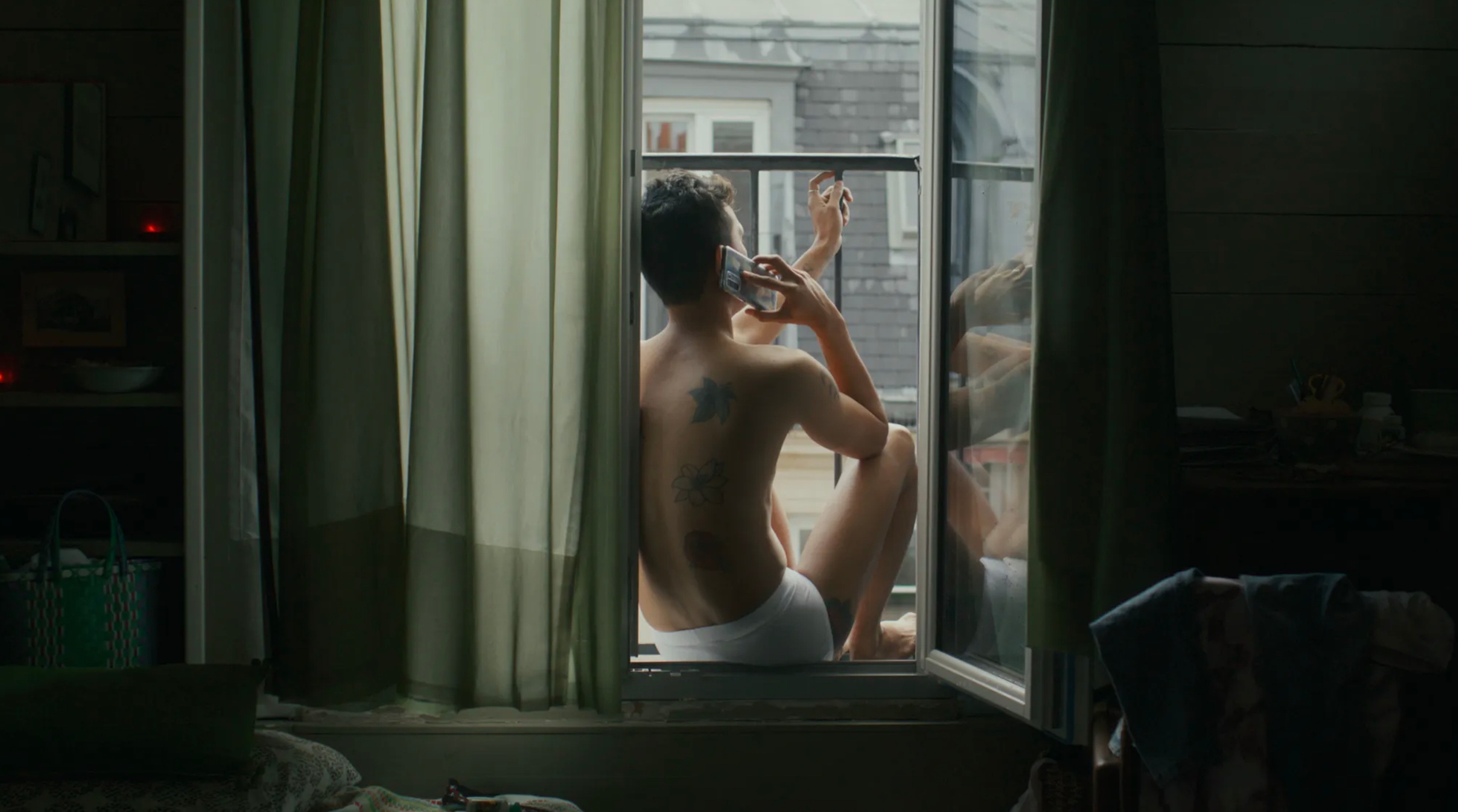 A man wearing underwear talks on the phone while sitting on a windowsill.