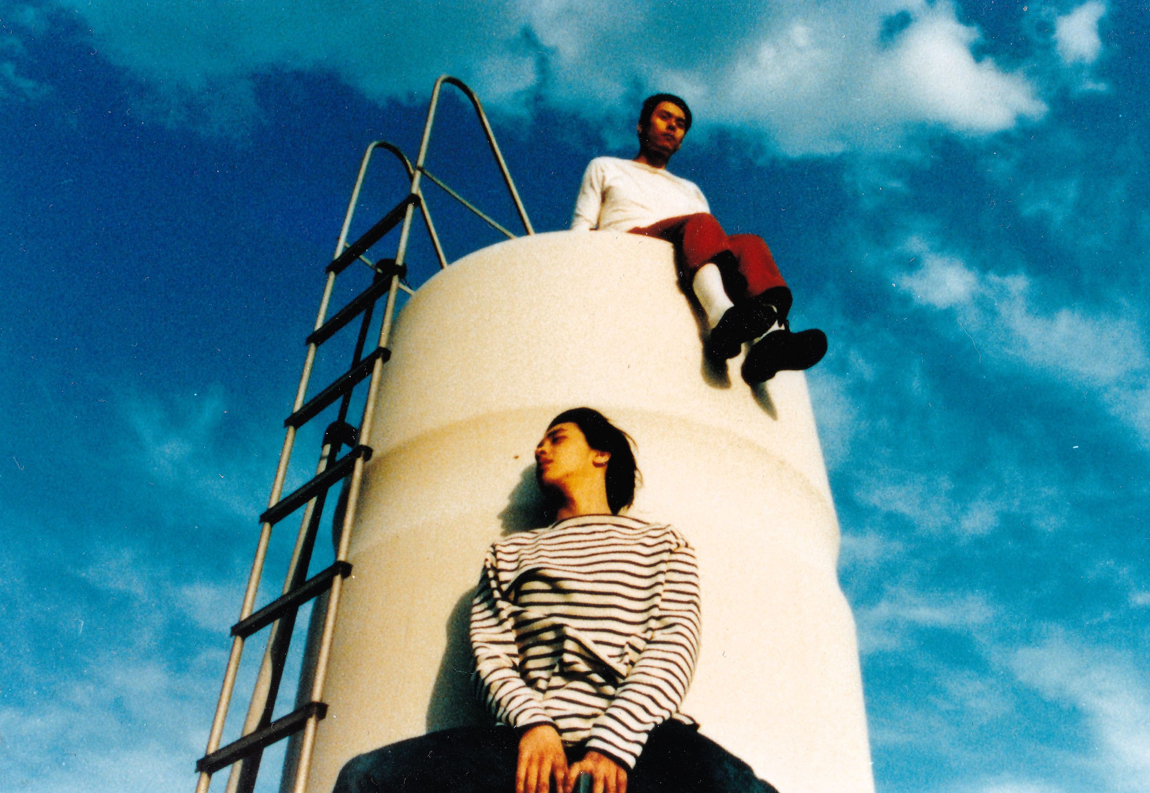 Two people sitting at different heights on a large white cylindrical structure beneath a vivid blue sky.