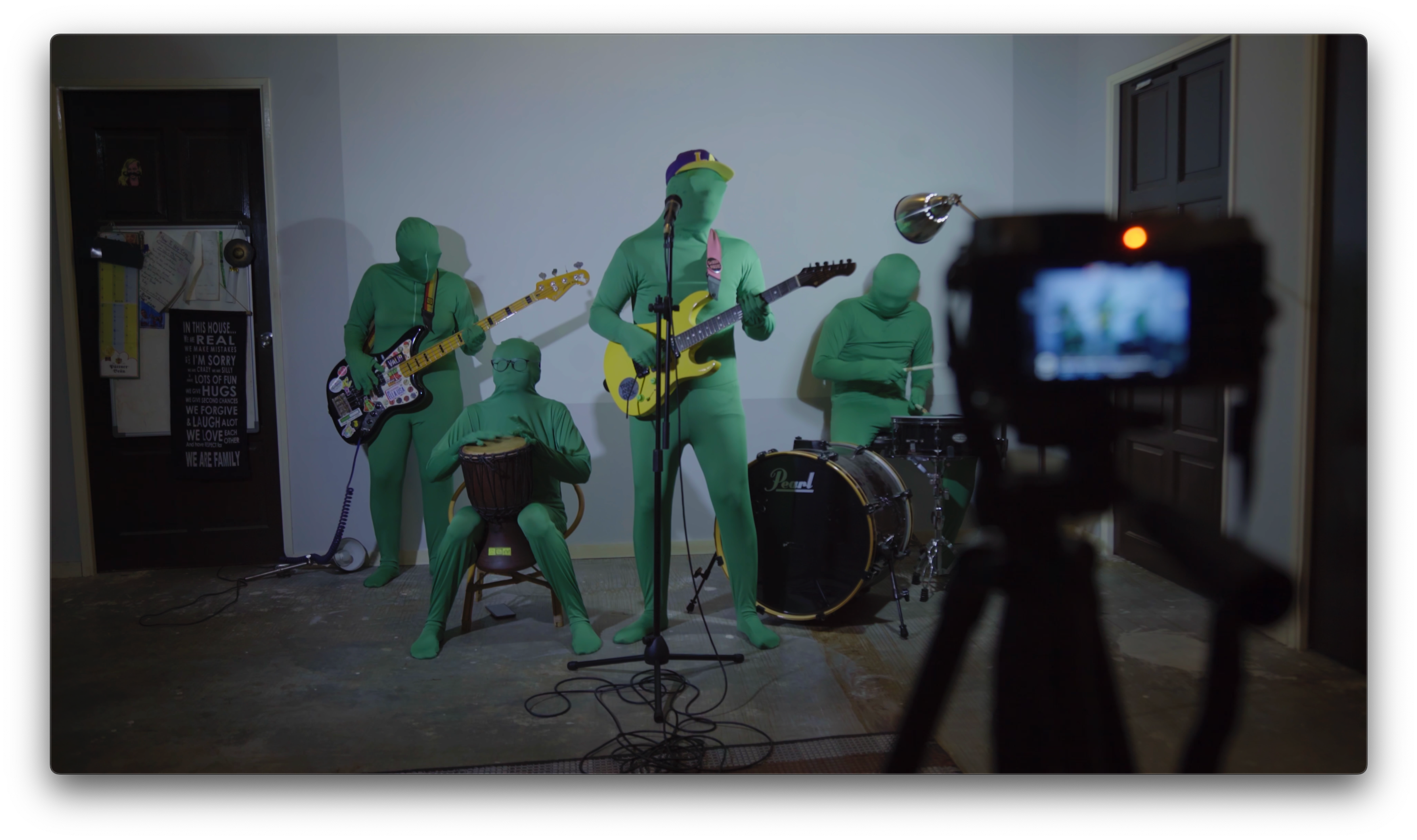 Four musicians in green bodysuits performing with guitars and drums while being filmed by a camera.
