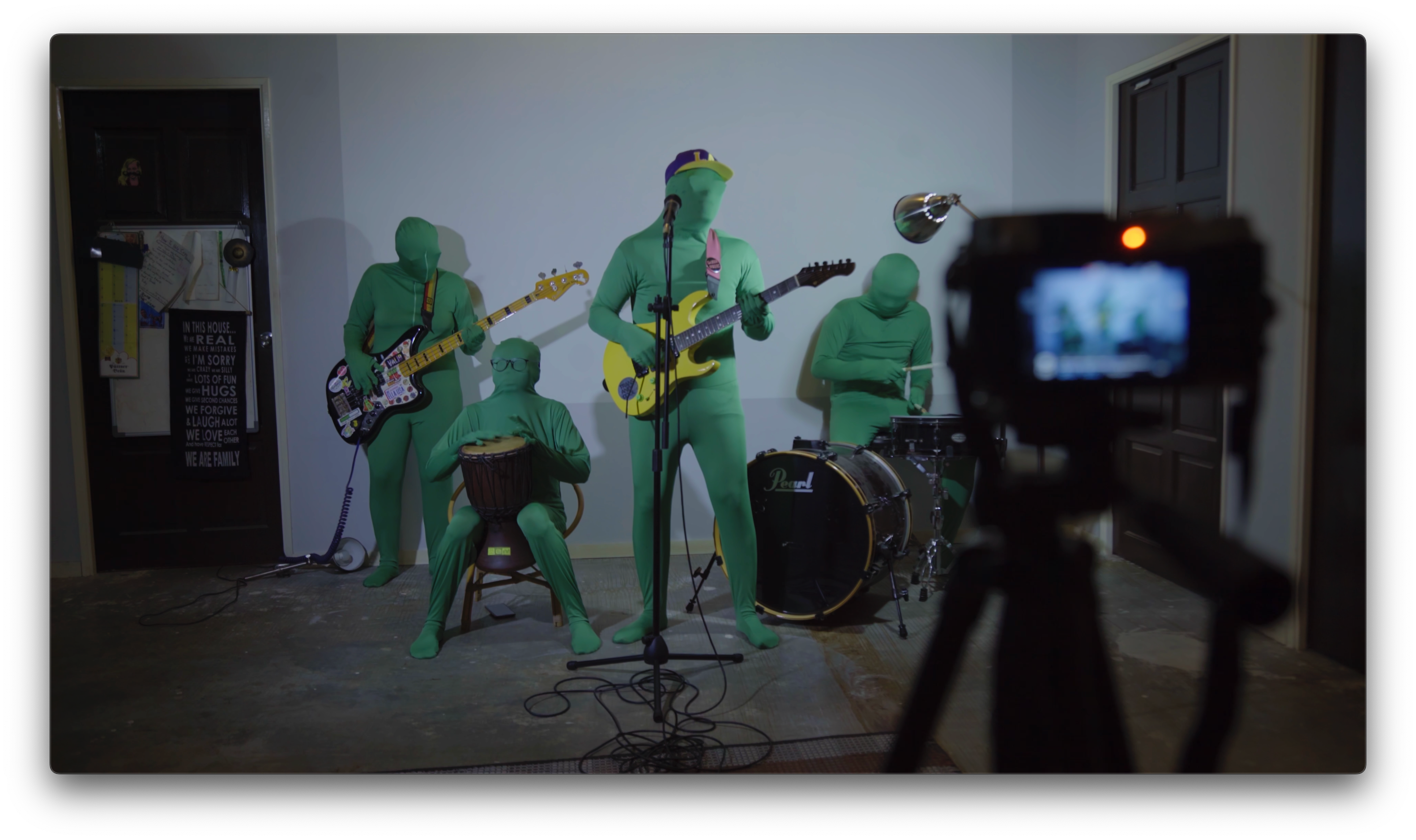 Four musicians in green bodysuits performing with guitars and drums while being filmed by a camera.