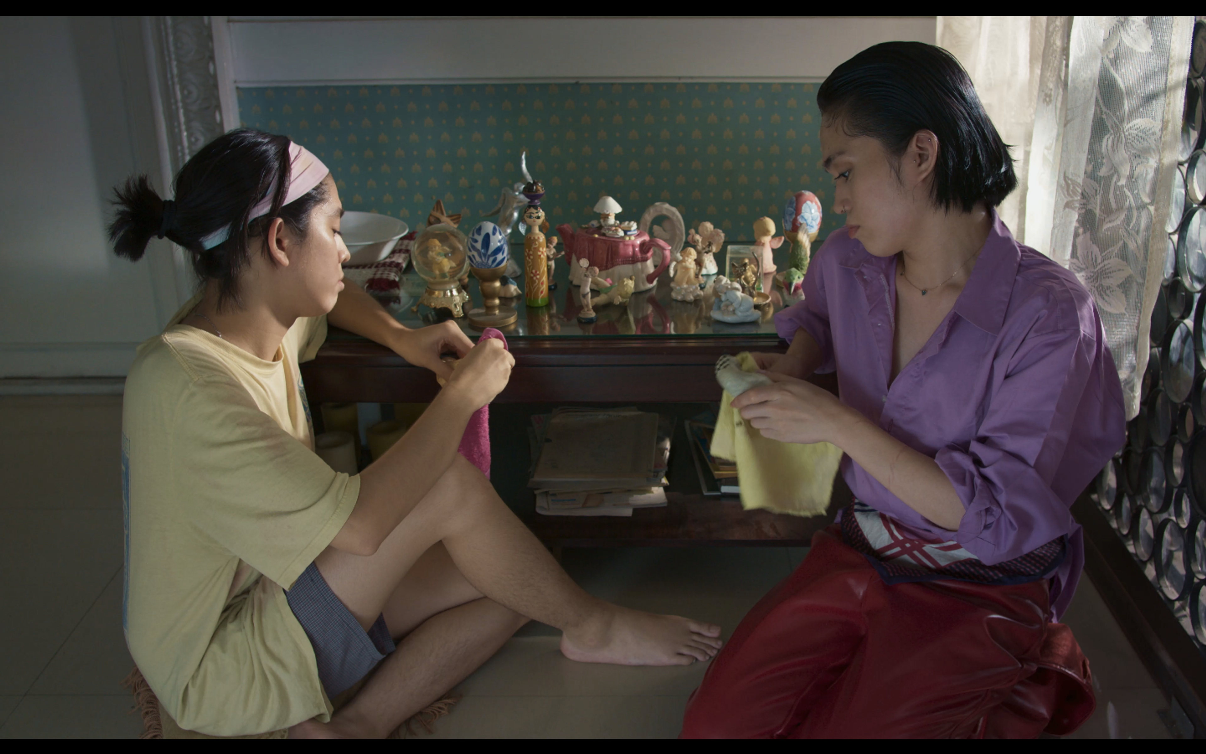 Two people sitting on the floor together, handling fabric beside a cabinet filled with small figurines.
