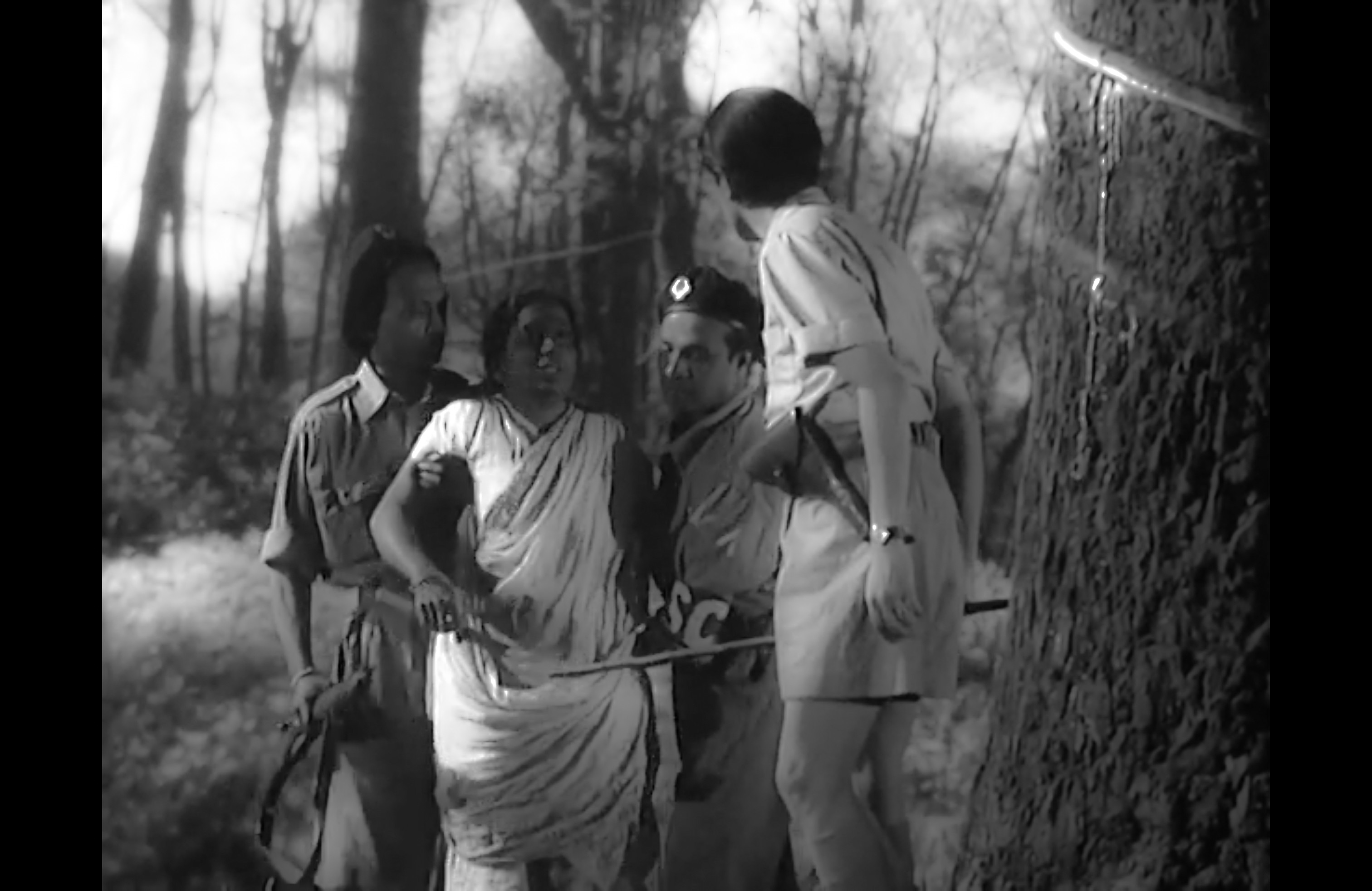 A black-and-white scene of four people gathered closely beside a large tree in a wooded setting.