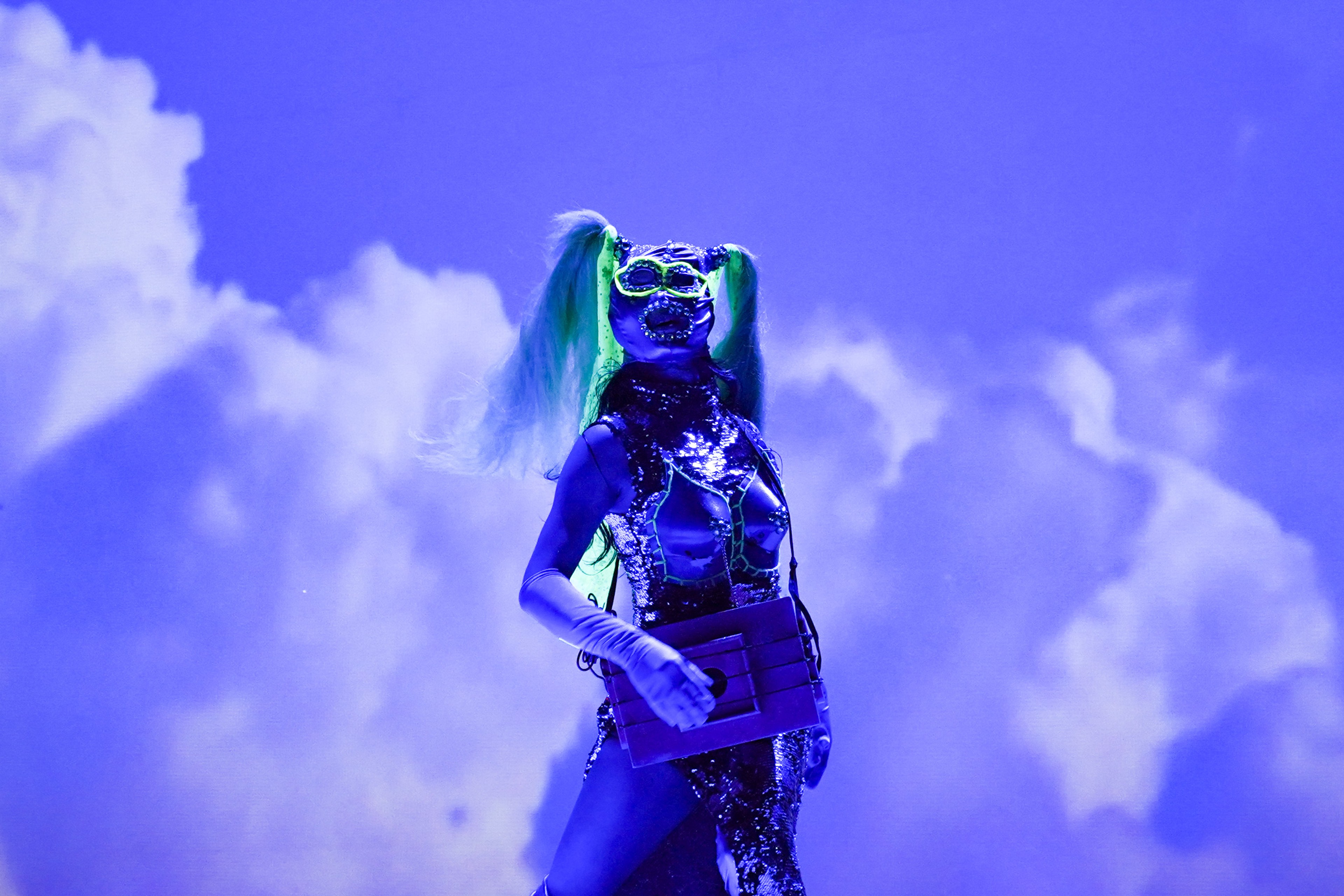 A performer in futuristic costume and goggles standing against a vivid blue, cloud-filled backdrop.