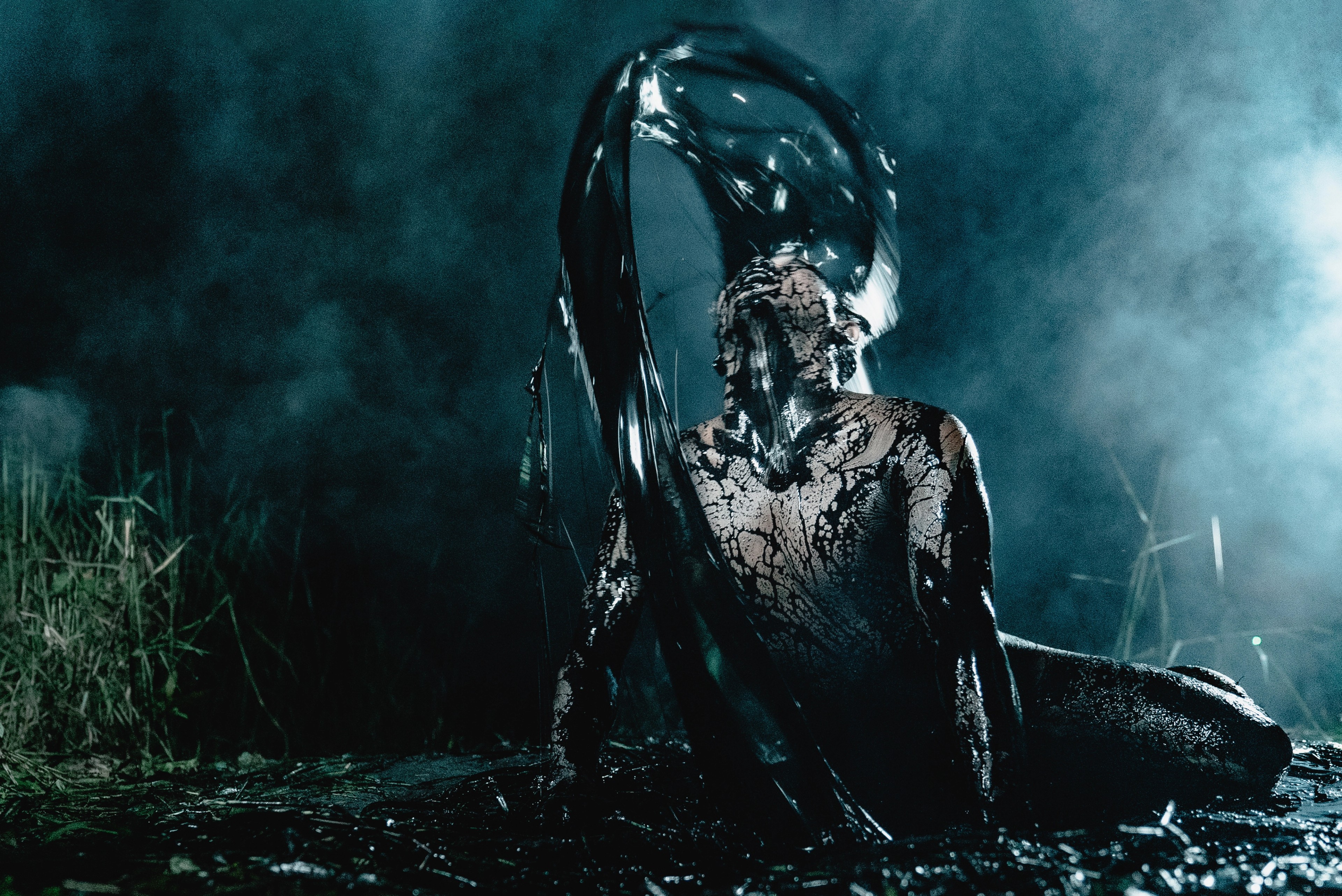 A figure covered in dark paint or mud sitting on wet ground beneath a sheet of black plastic in a smoky nighttime setting.