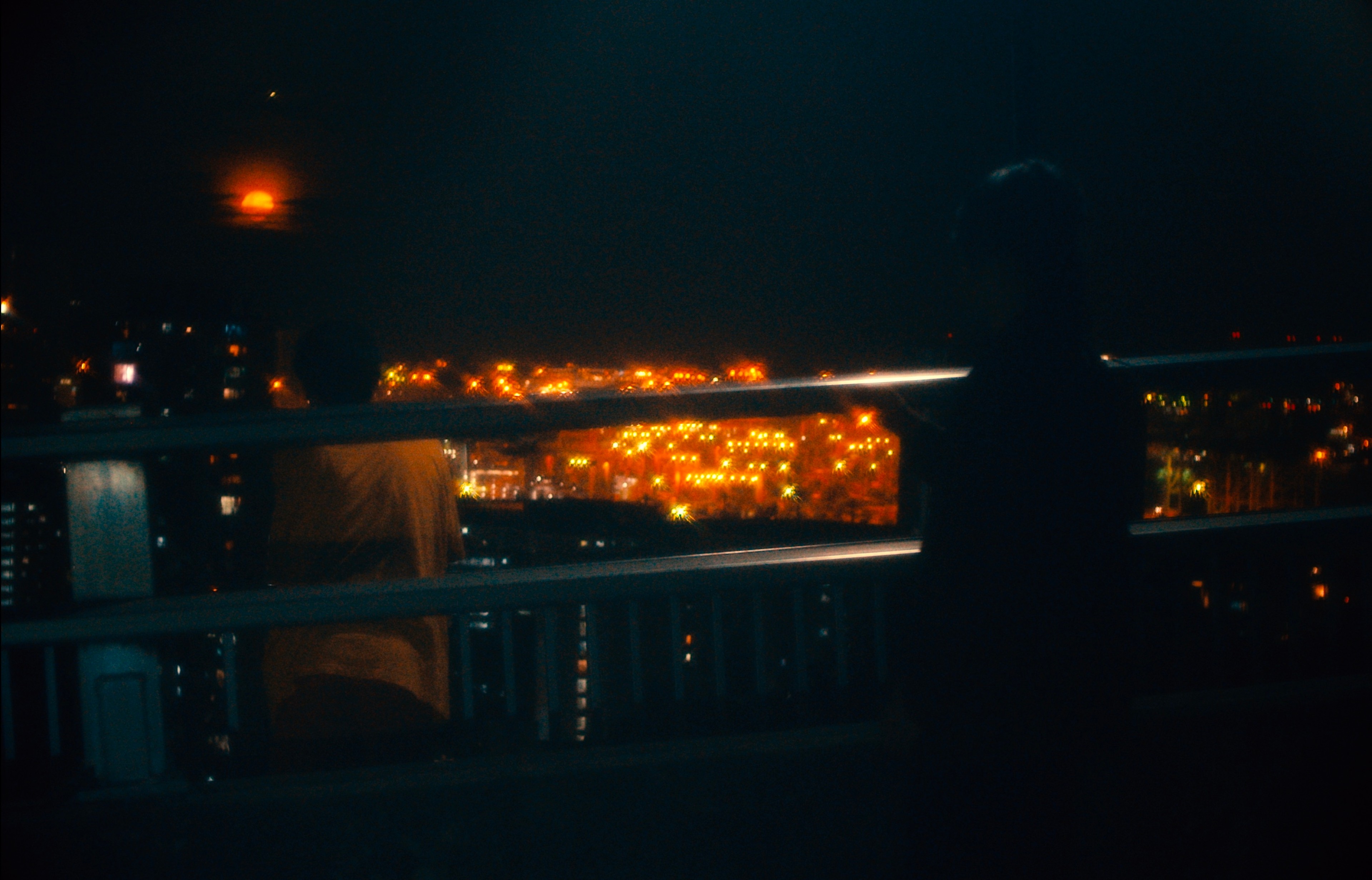 A silhouetted figure standing on a balcony at night, overlooking a city glowing with orange lights.