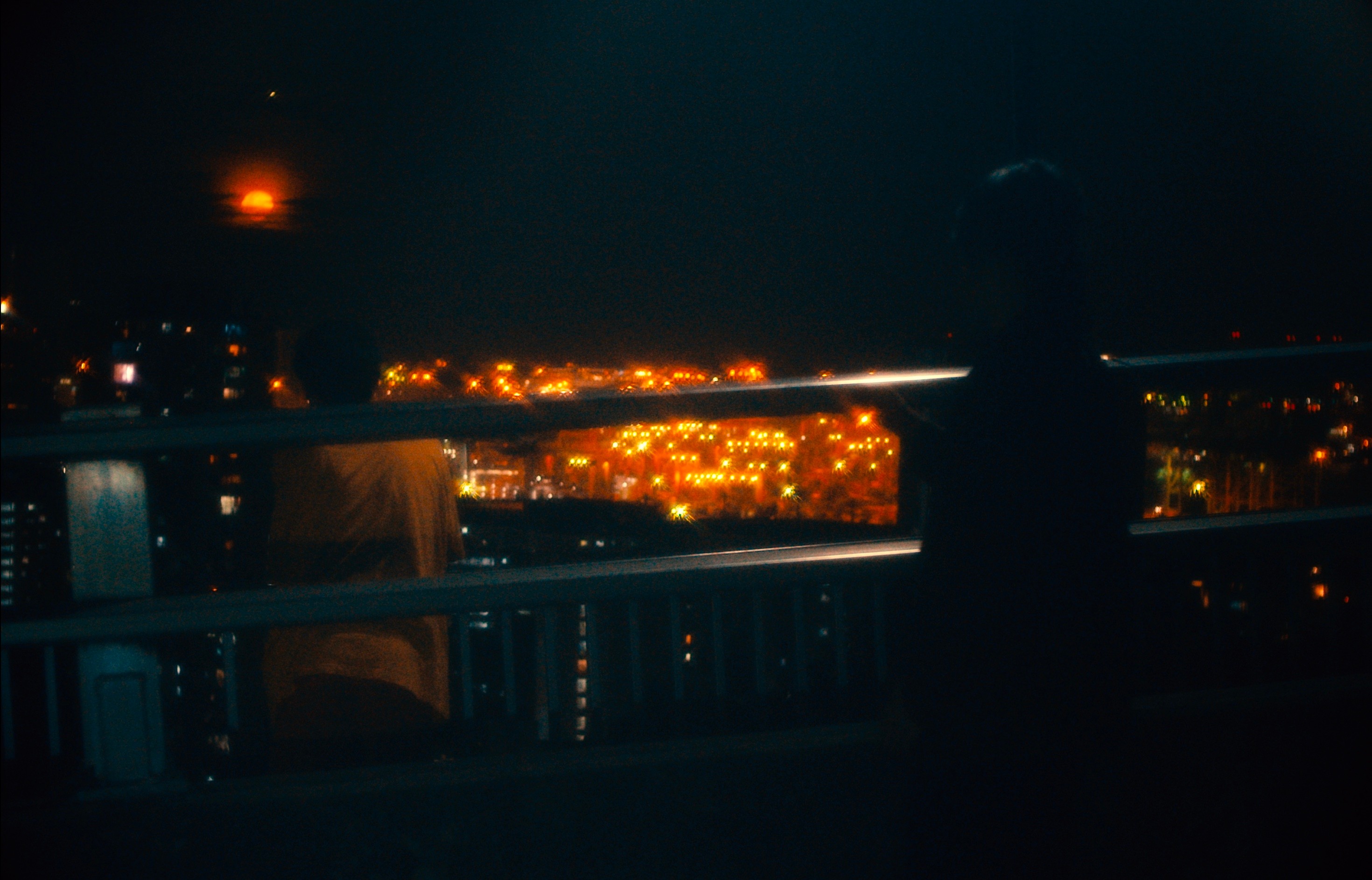 A silhouetted figure standing on a balcony at night, overlooking a city glowing with orange lights.