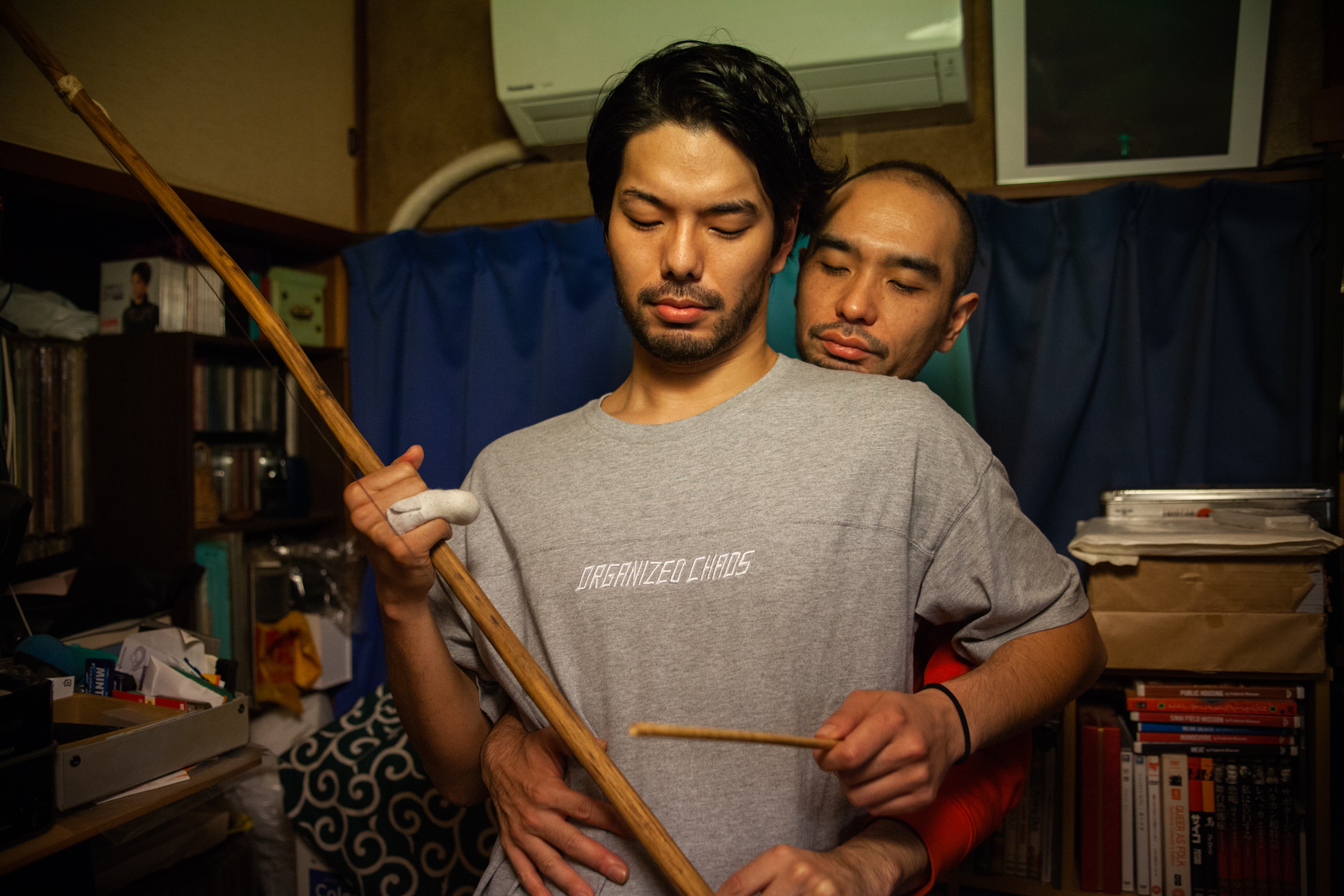 Two men standing closely together in a cluttered room, one holding a long wooden pole while the other embraces him from behind.