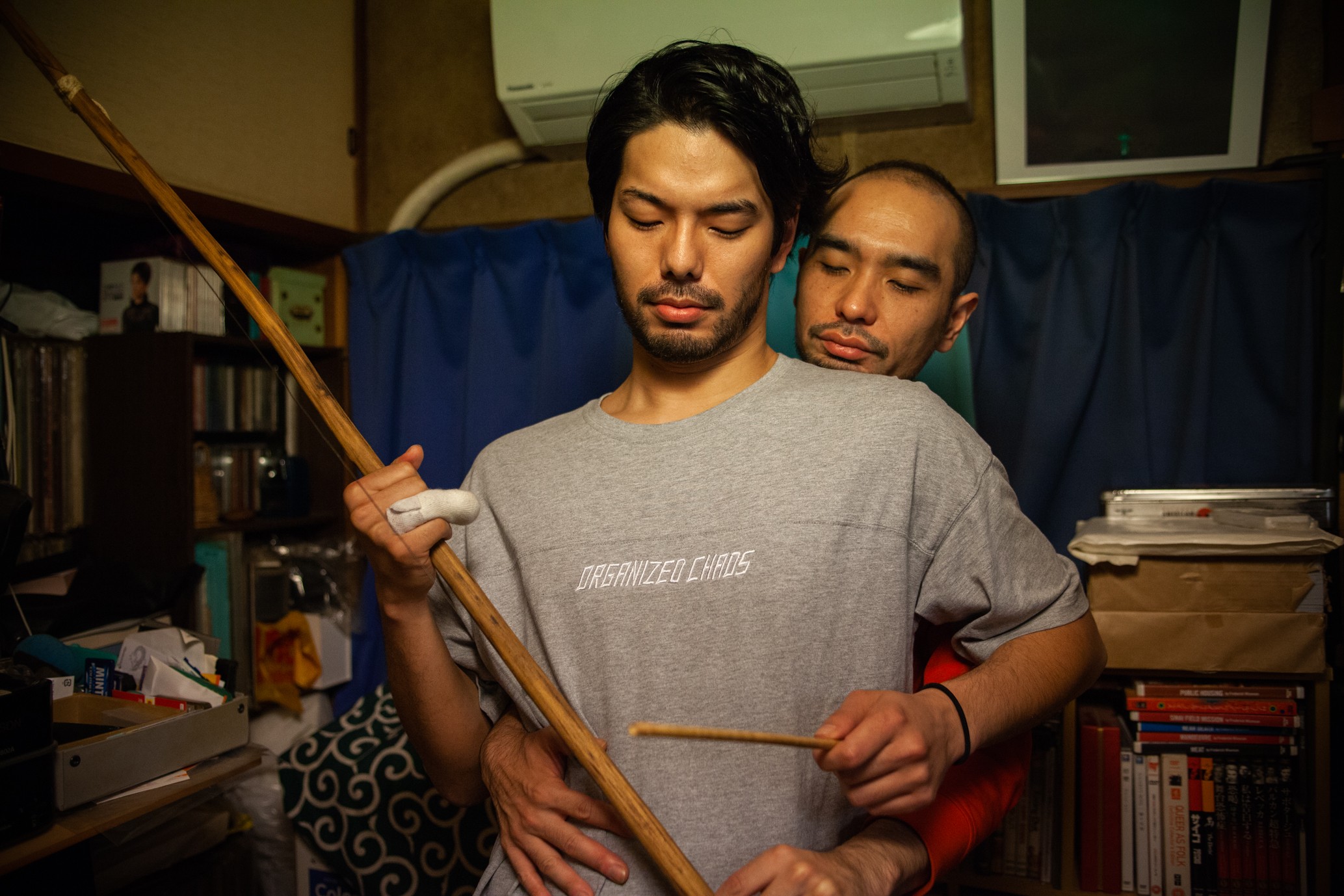 Two men standing closely together in a cluttered room, one holding a long wooden pole while the other embraces him from behind.