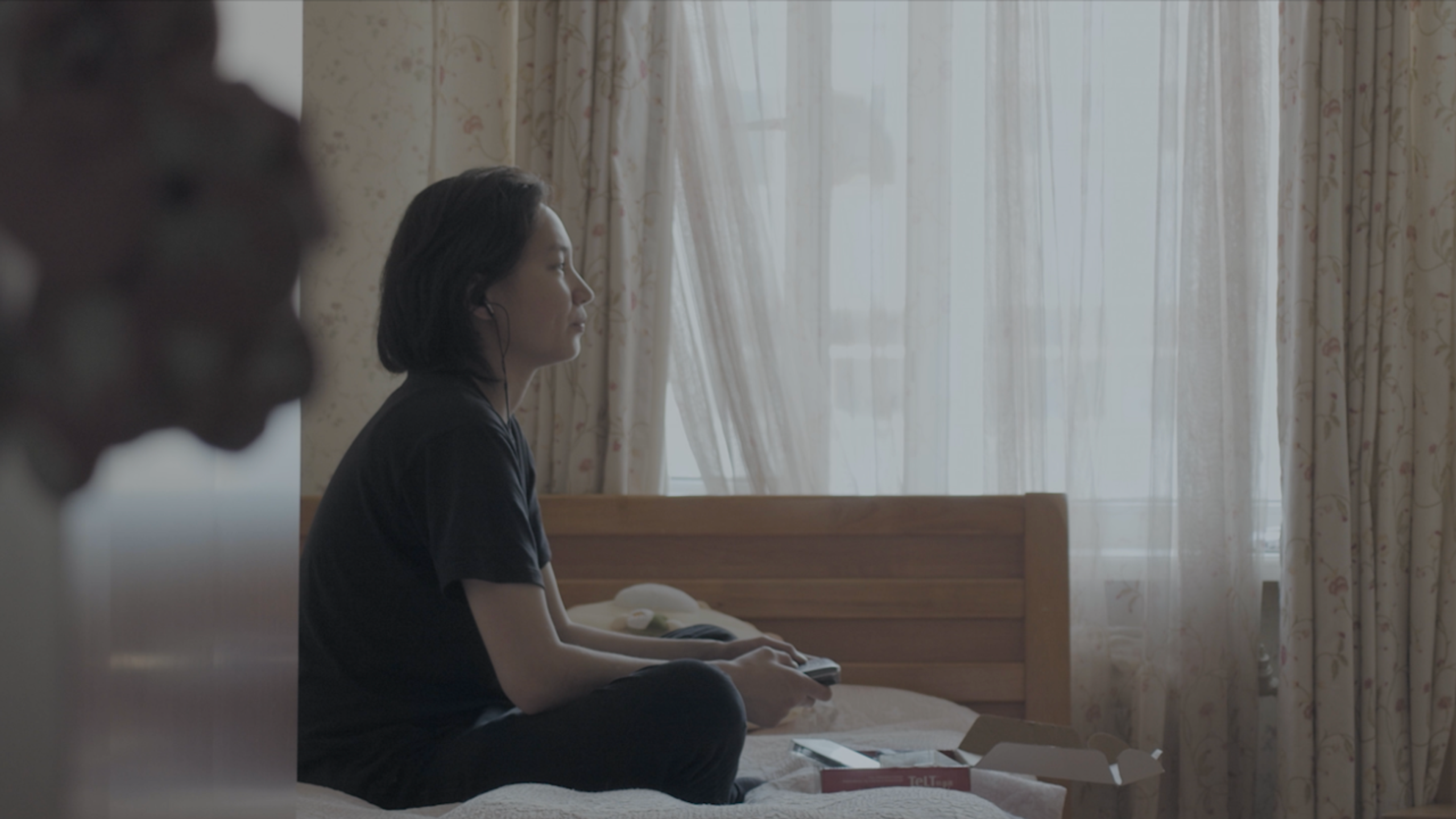 A woman sitting cross-legged on a bed, looking toward a softly lit window in a quiet bedroom.