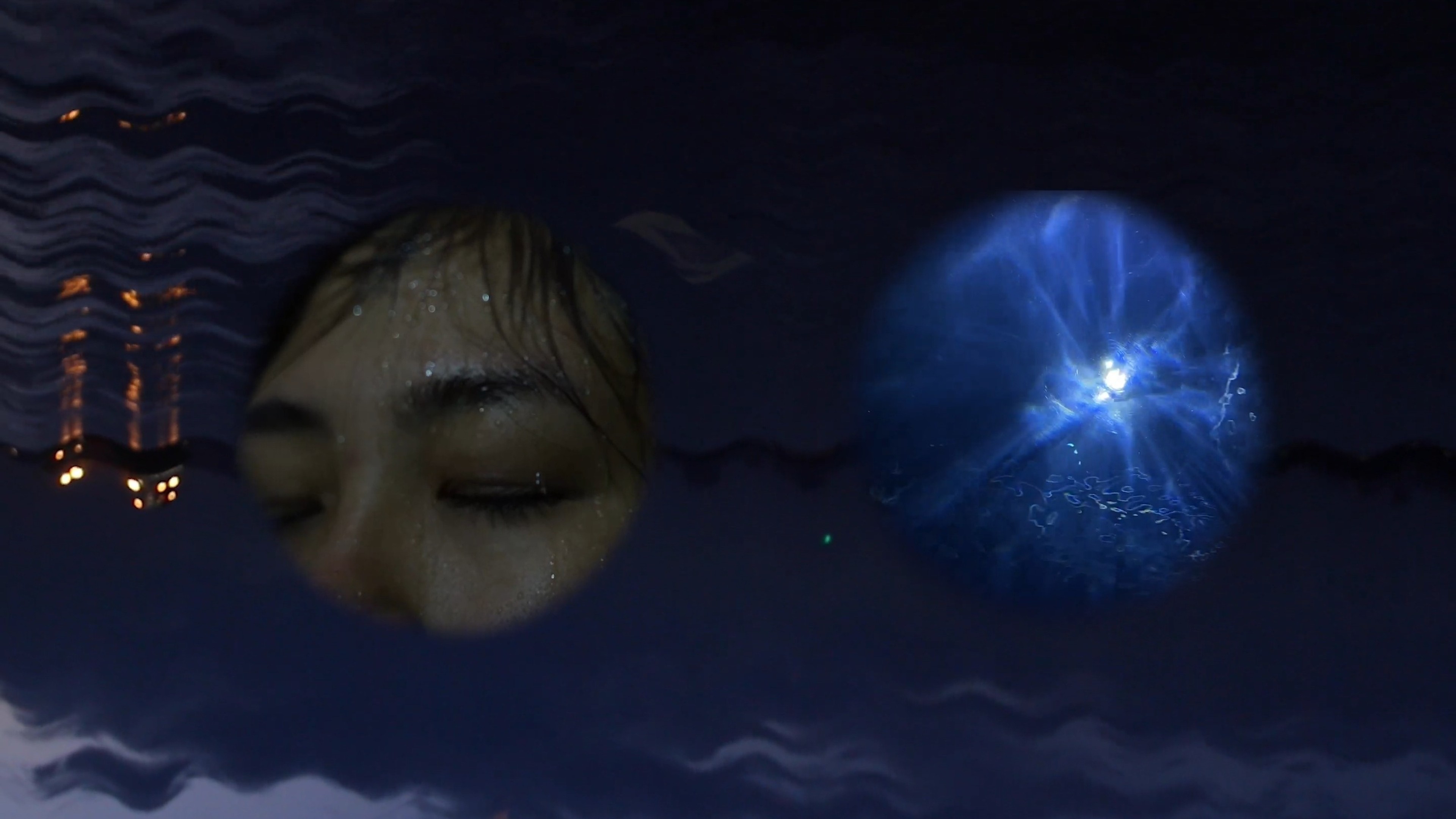 A submerged face with closed eyes beside a glowing blue jellyfish-like form in dark water.