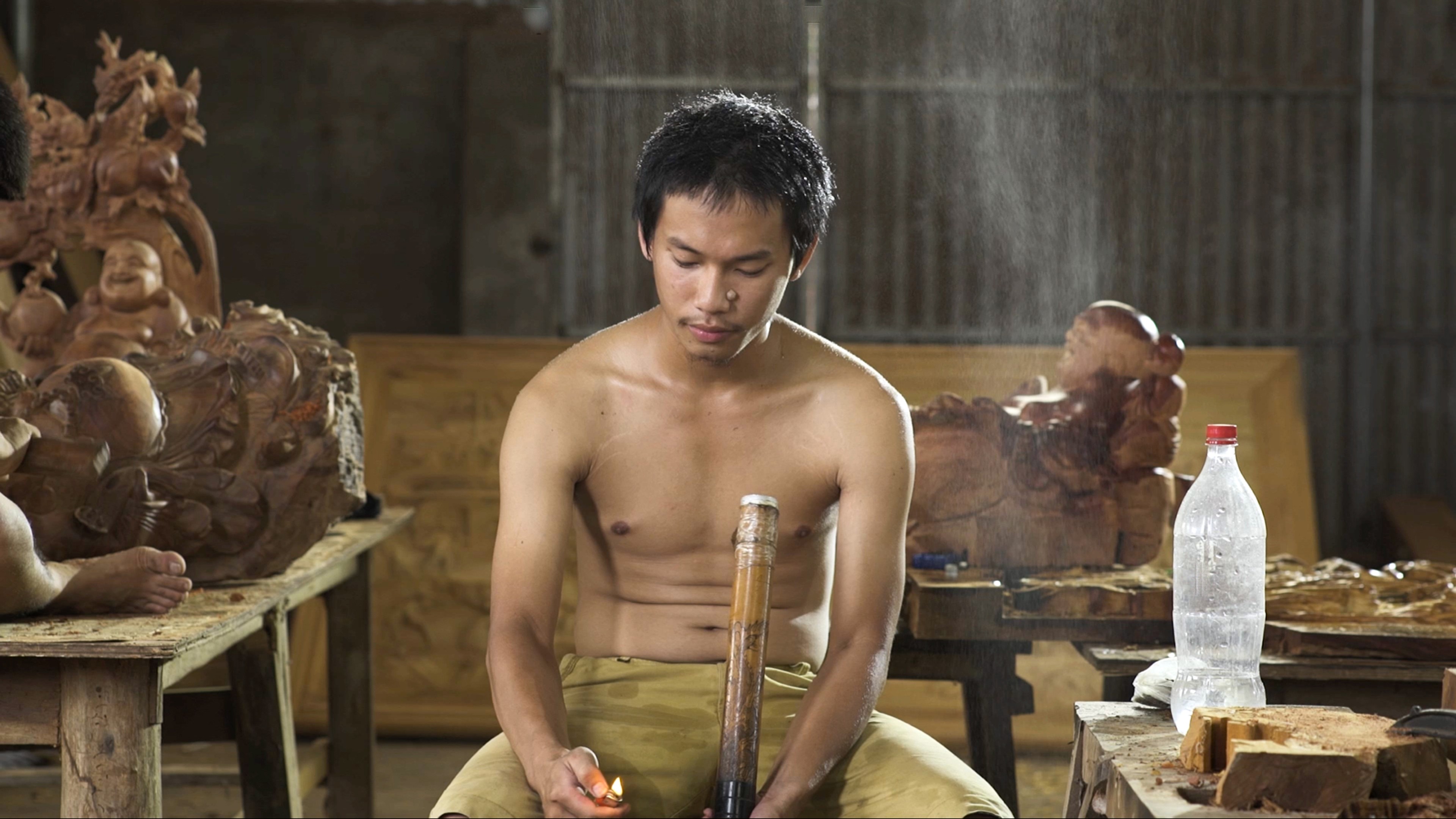 A topless man holds a lit lighter surrounded by wooden carvings.
