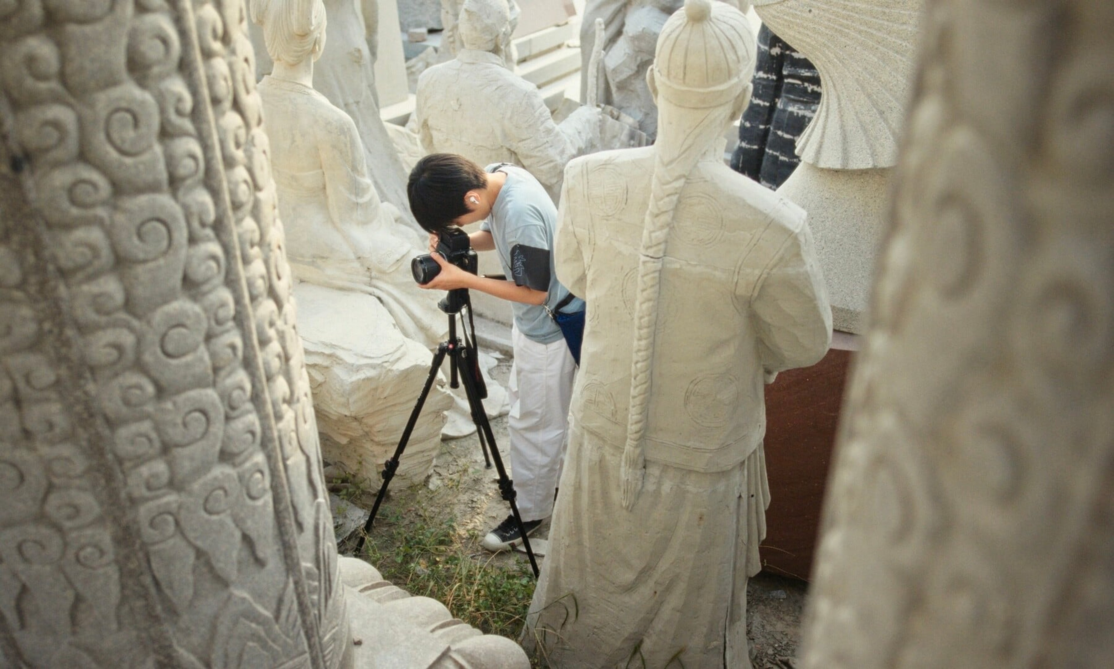 A person holds a camera in the midst of several white statues.