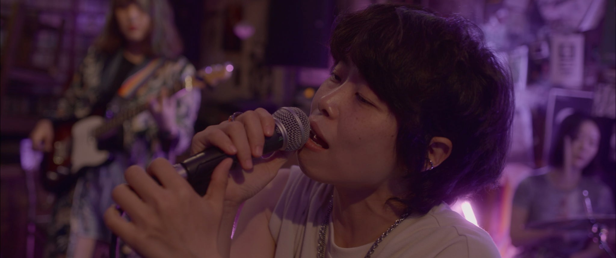 A singer performing into a microphone under purple stage lighting, with band members playing behind them.