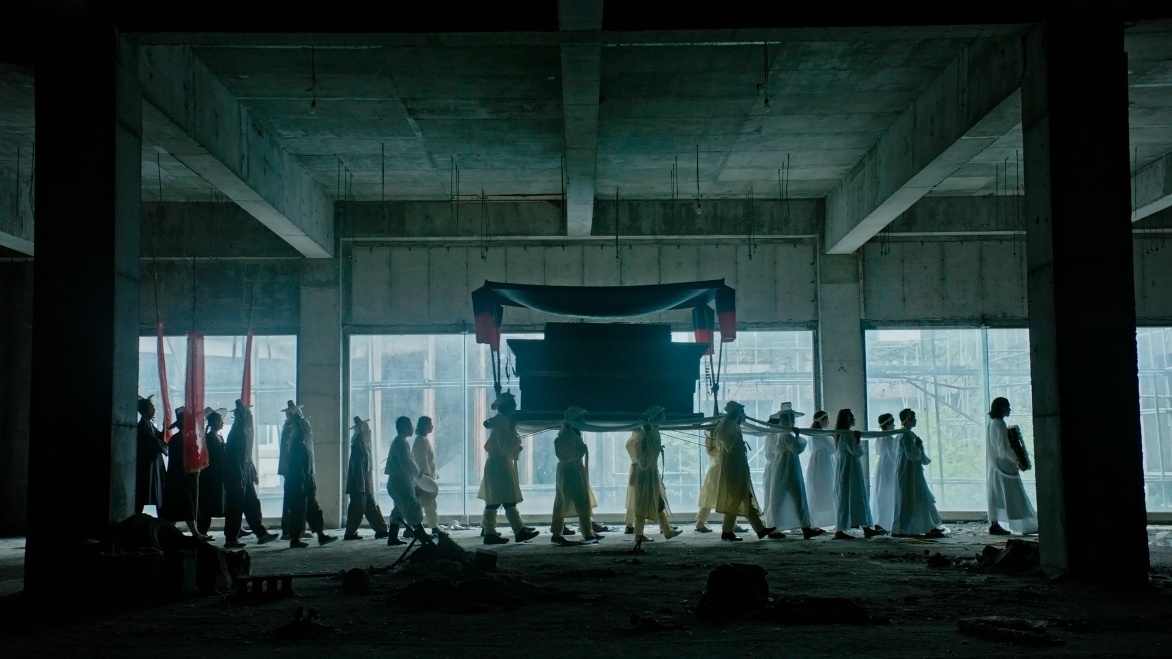 A procession of people holding a coffin walk through an abandoned building.