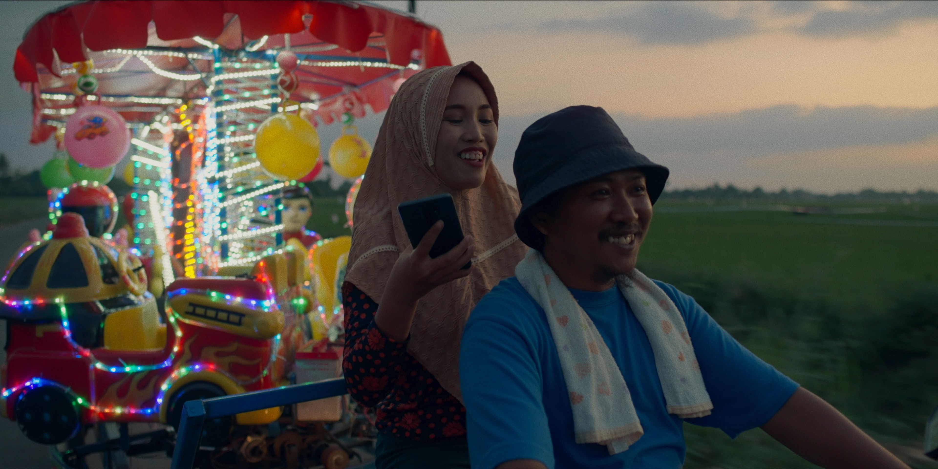 A smiling man and woman riding together beside a brightly lit fairground ride at dusk.