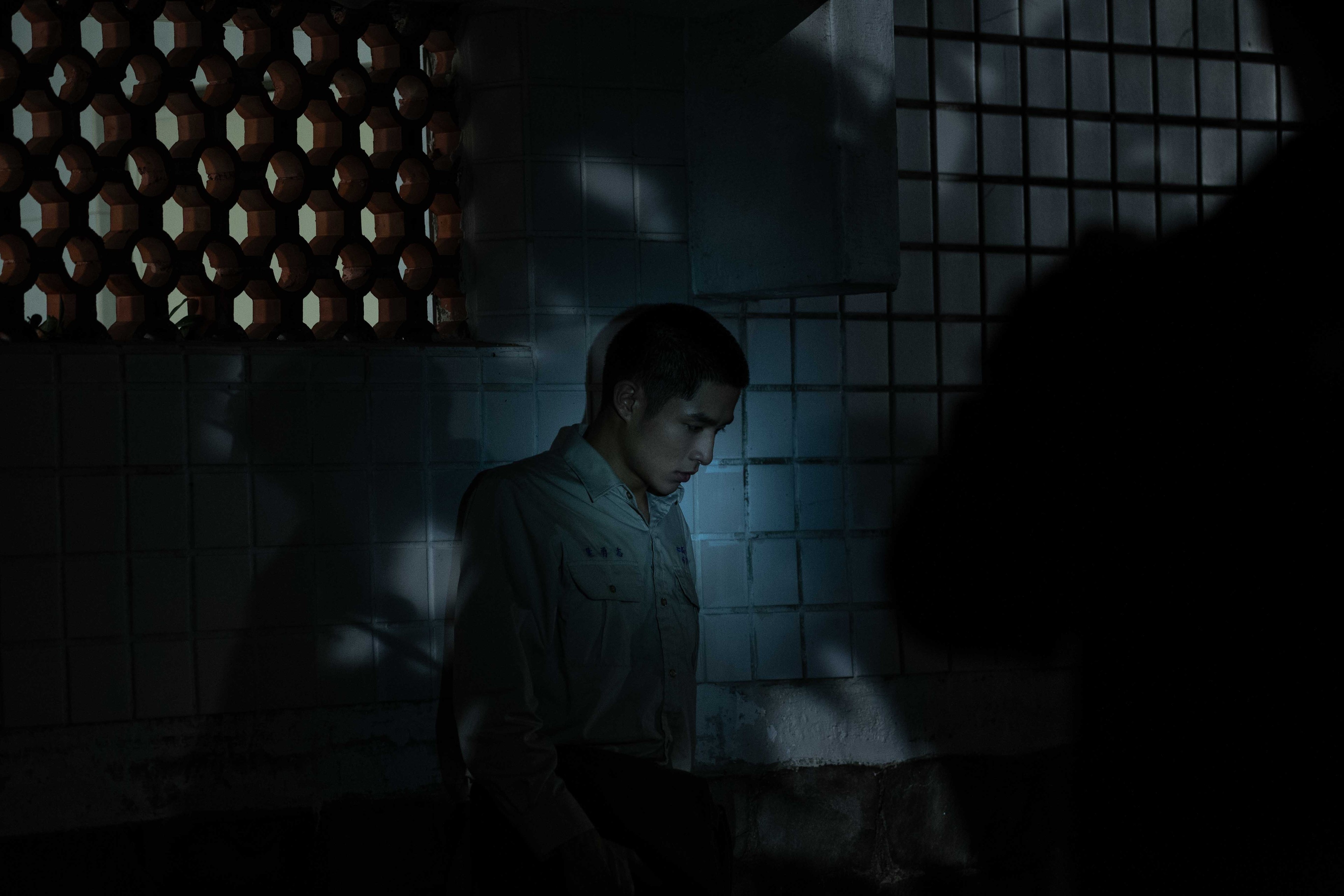 A man stands against a tiled wall in a dark room.