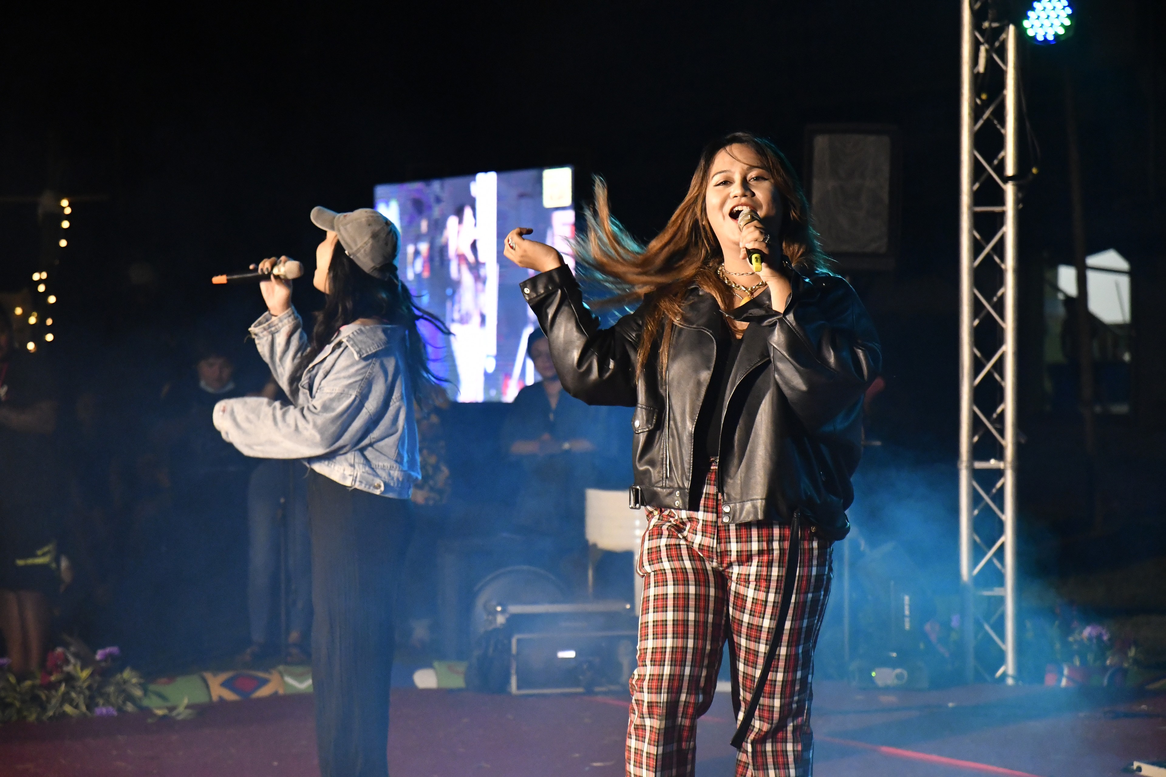 Two performers singing on an outdoor stage at night, one gesturing toward the audience while holding a microphone.