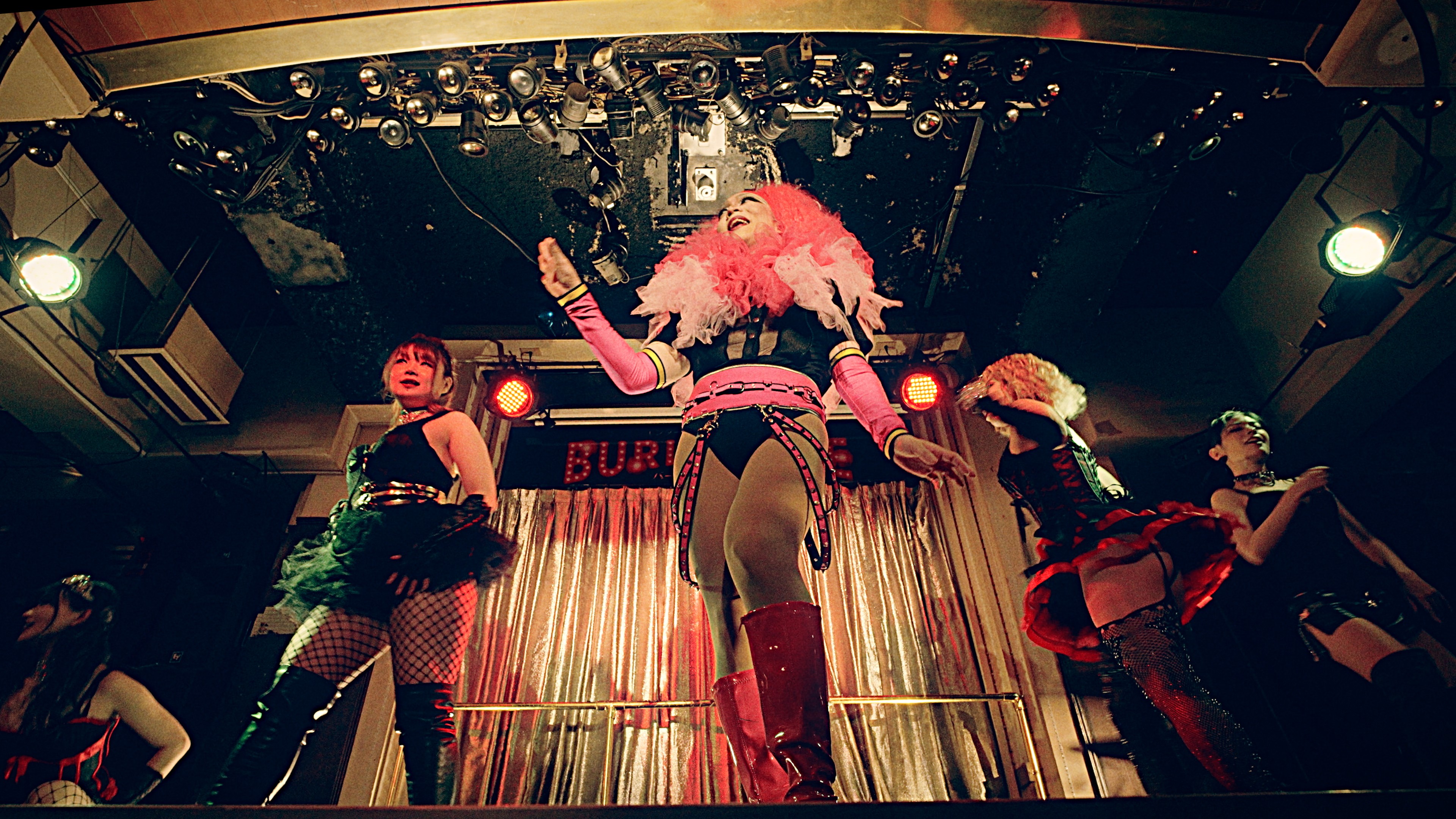 A group of performers in colourful costumes dancing onstage under bright club lights.