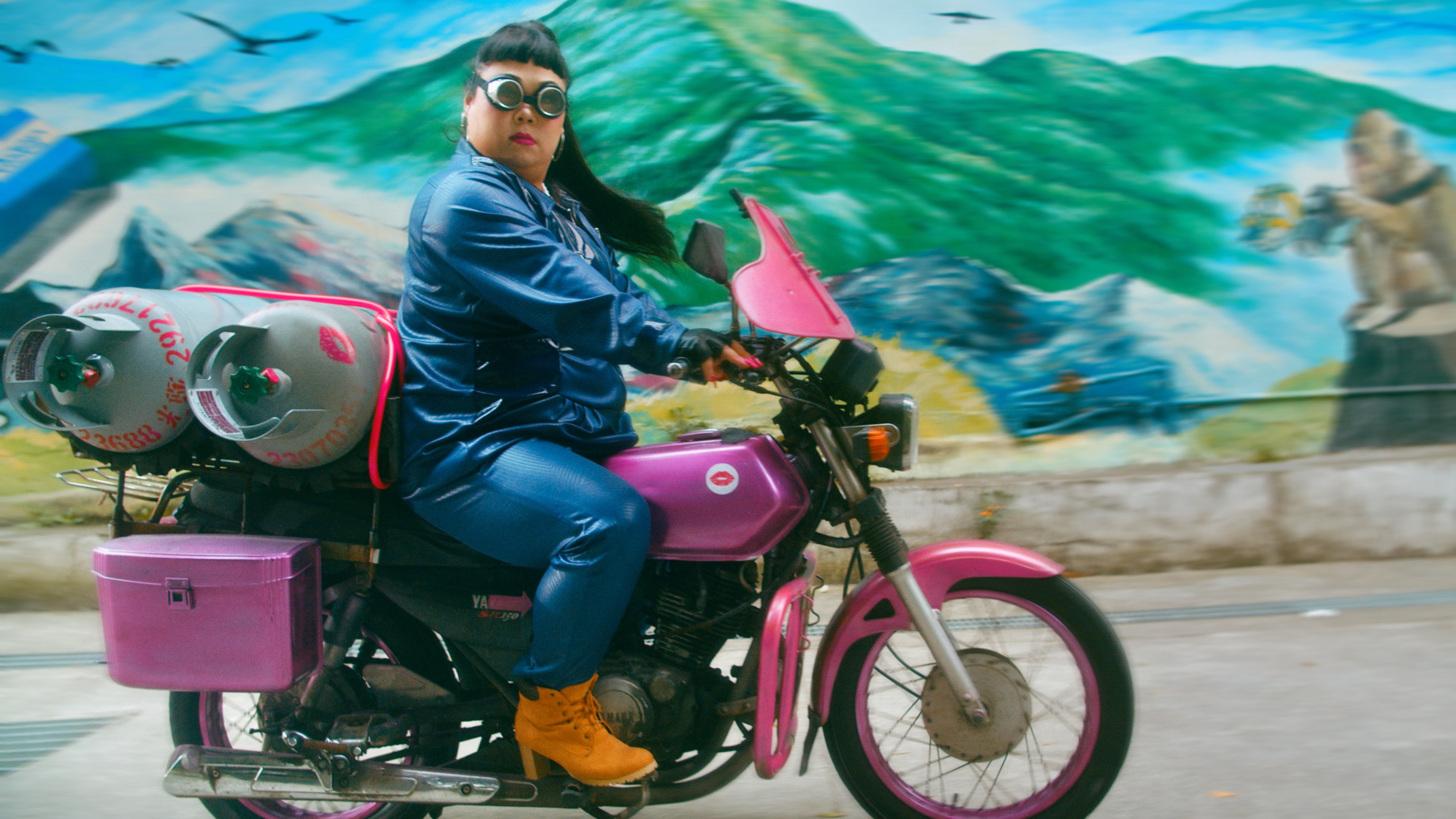 A person wearing goggles and a blue jumpsuit riding a bright pink motorcycle past a colourful mural.