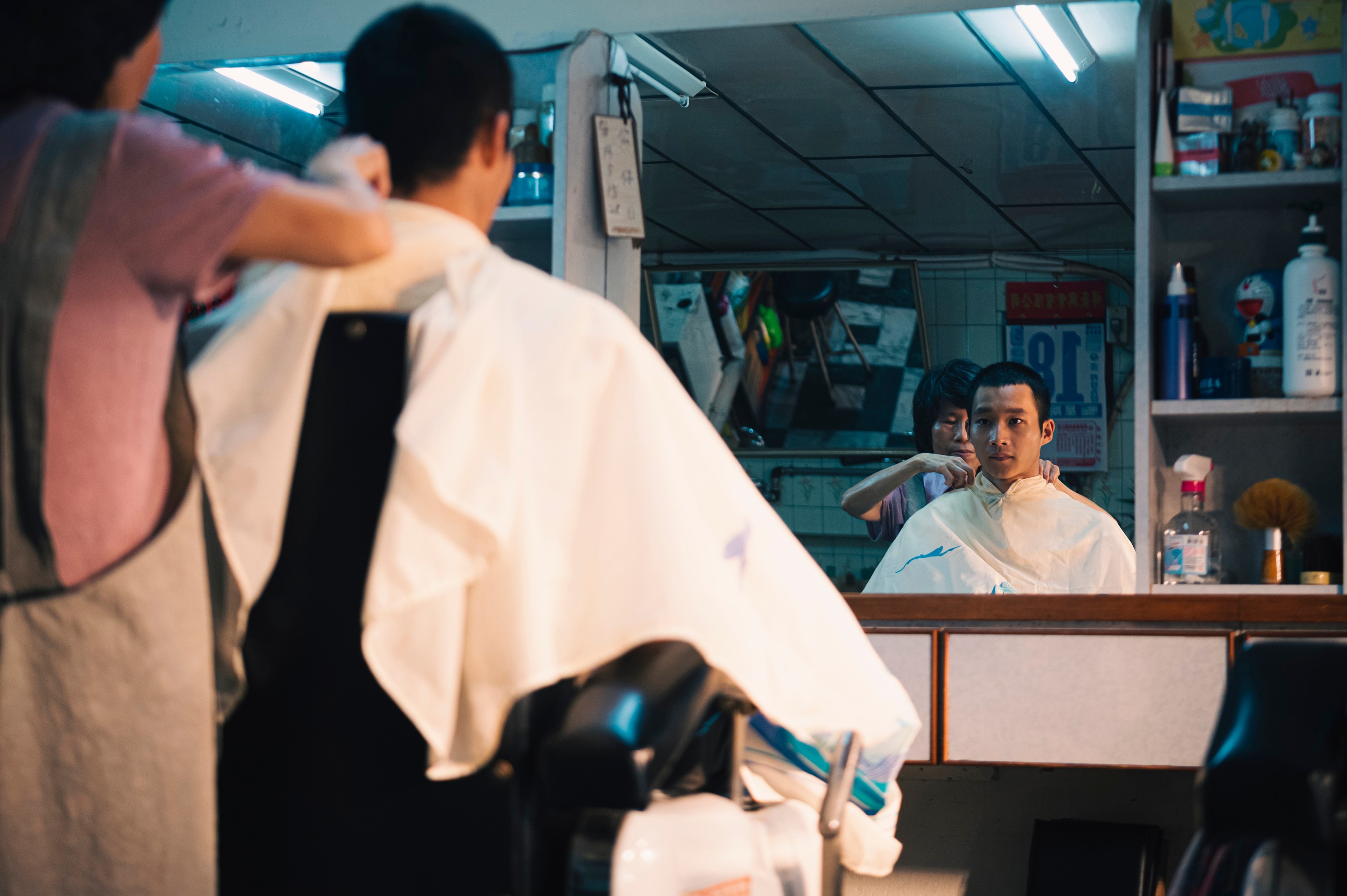 A man receiving a haircut in a small barbershop, seen reflected in the mirror.