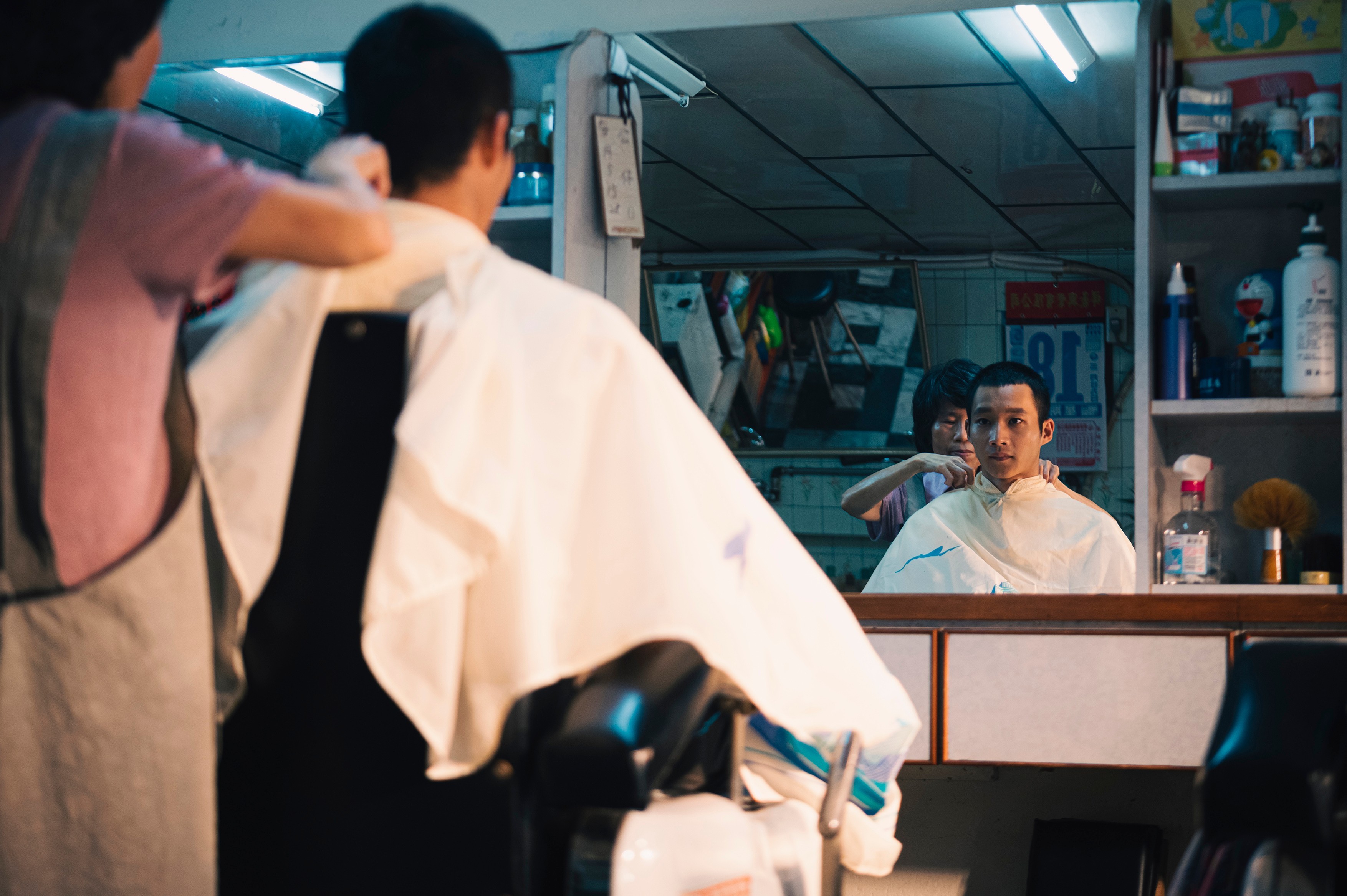 A man receiving a haircut in a small barbershop, seen reflected in the mirror.