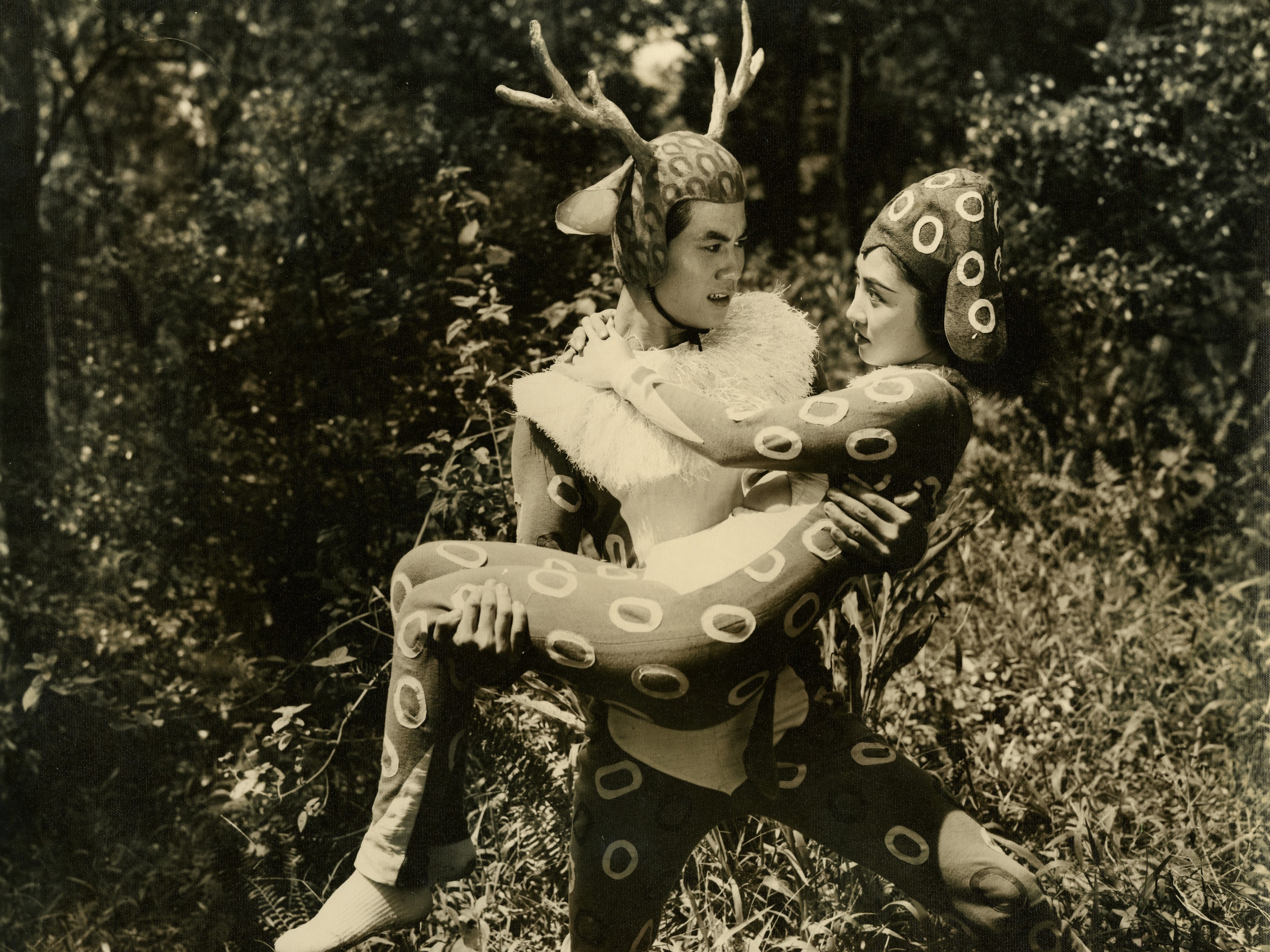 A man wearing antlers holds a woman in their arms in a wooded setting.