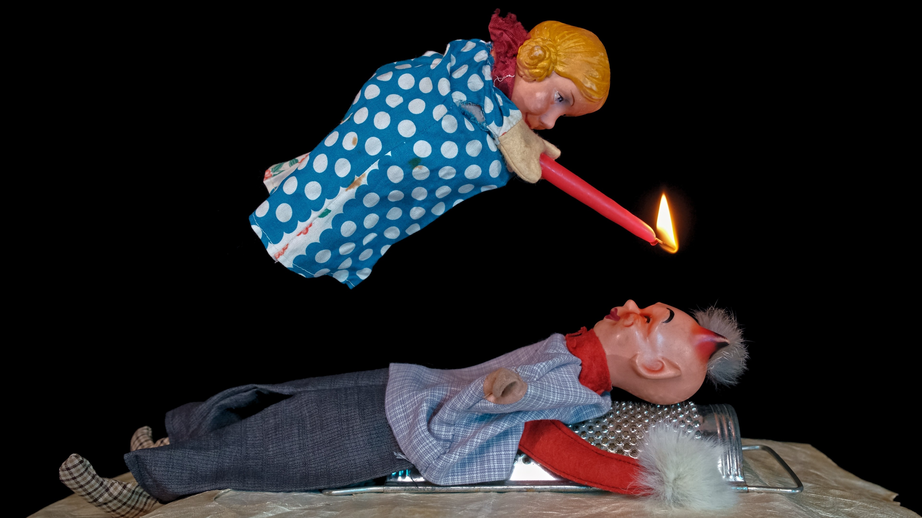 A puppet holding a lit candle hovers above another puppet.