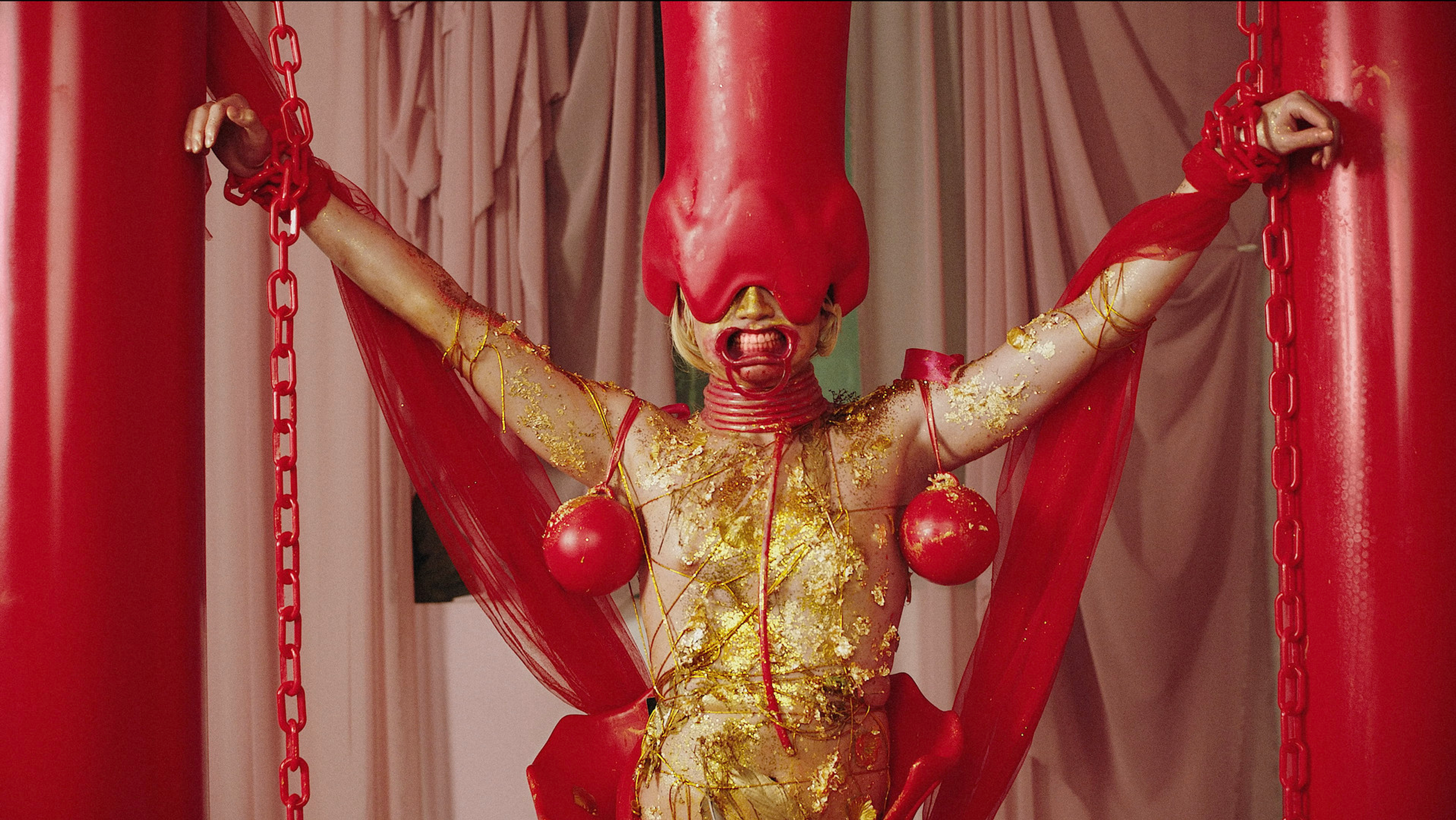 A performer in an elaborate red-and-gold costume posed with arms outstretched between red chains and columns.