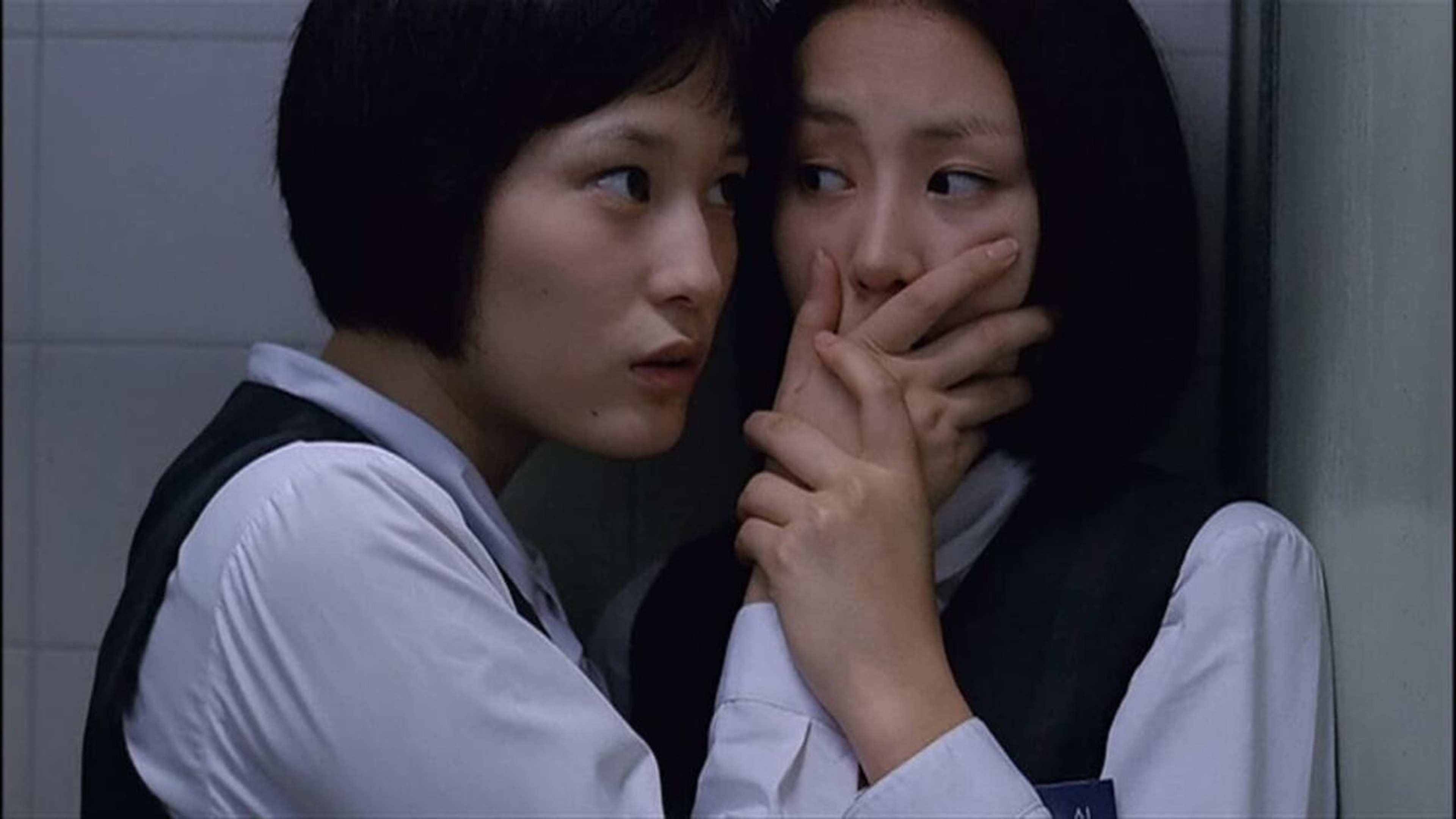 Two young women in school uniforms standing close together, one covering the other’s mouth with her hand.