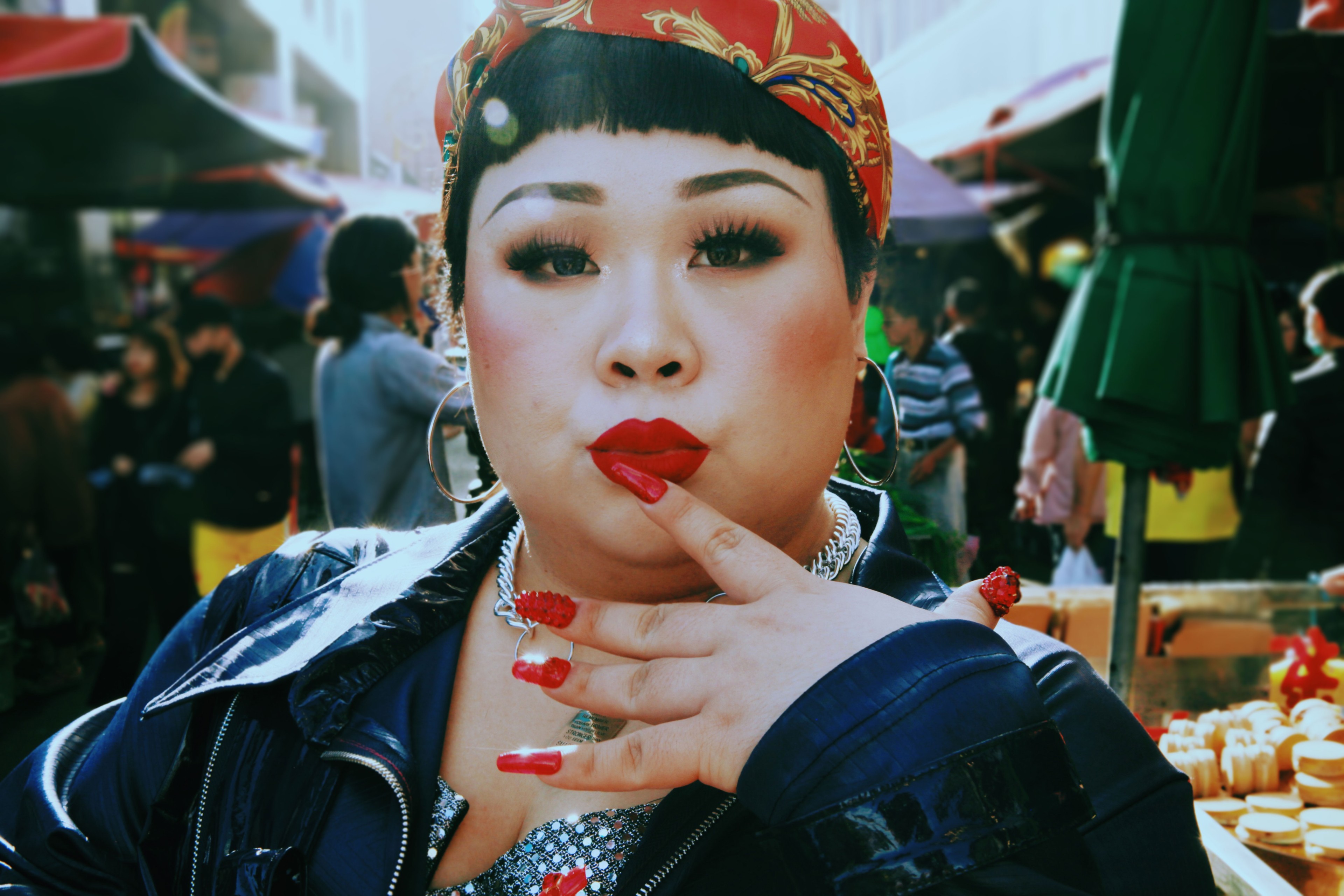 A person in bold makeup, a patterned headscarf and black jacket posing at a busy outdoor market.