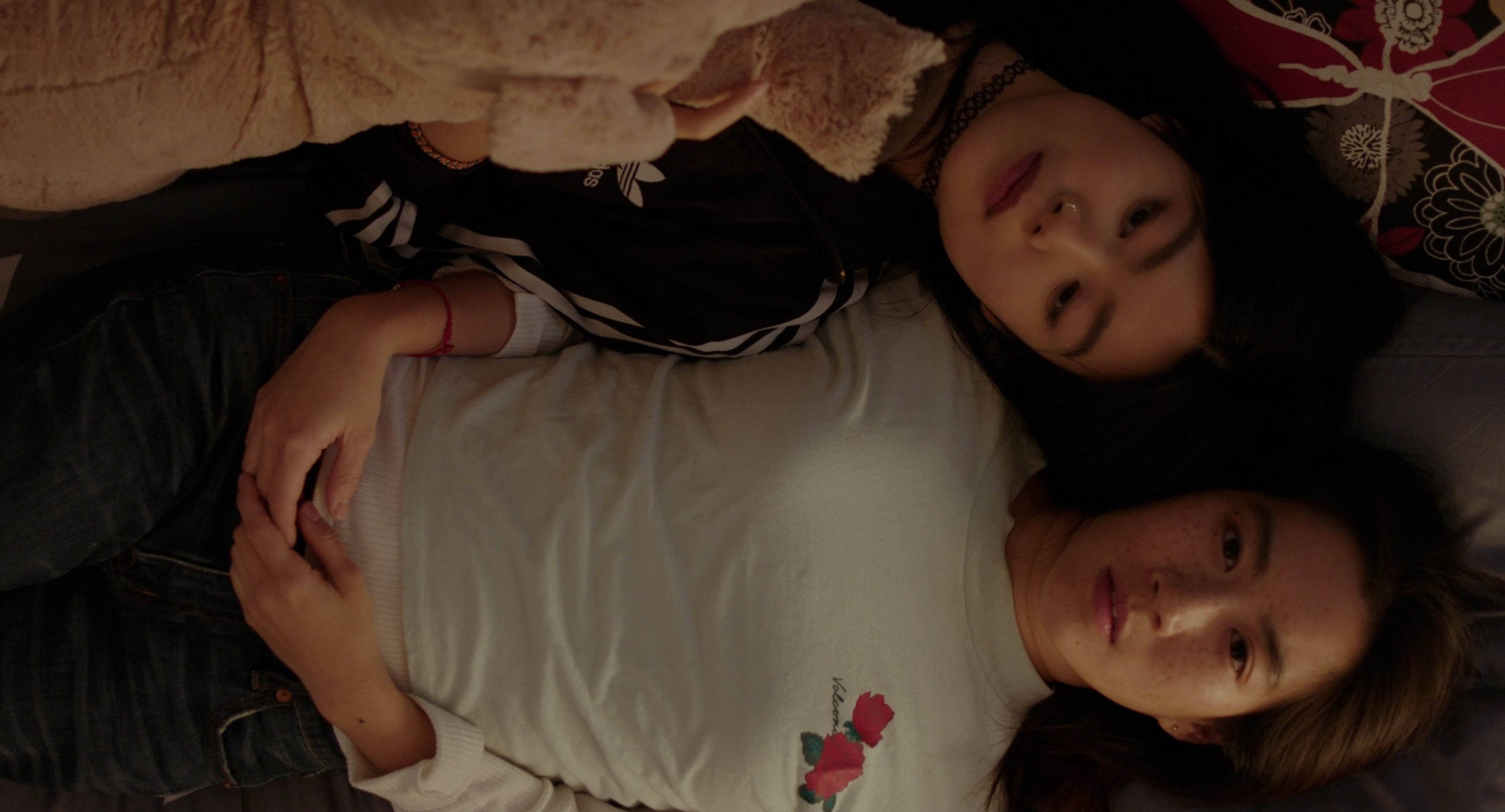 Two women lying closely together on a bed, looking upward in a softly lit room.