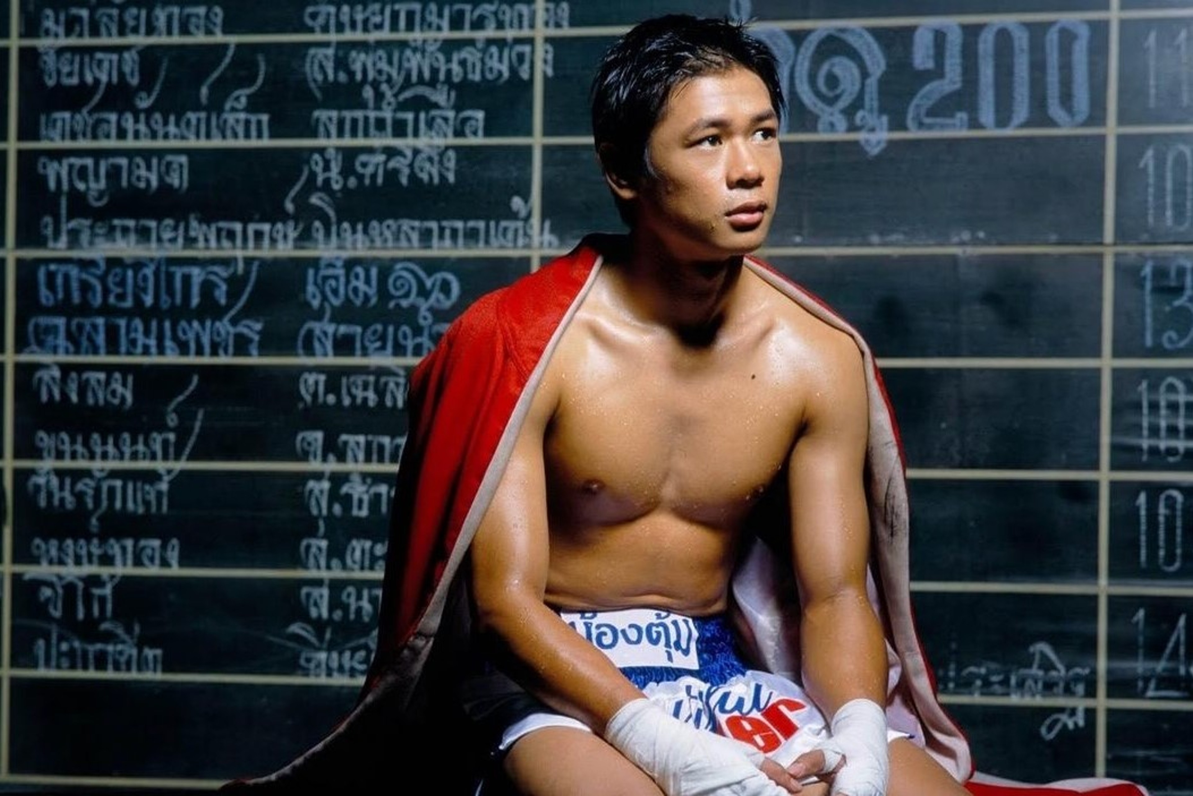 A shirtless boxer with wrapped hands sitting beneath a red robe in front of a handwritten scoreboard.