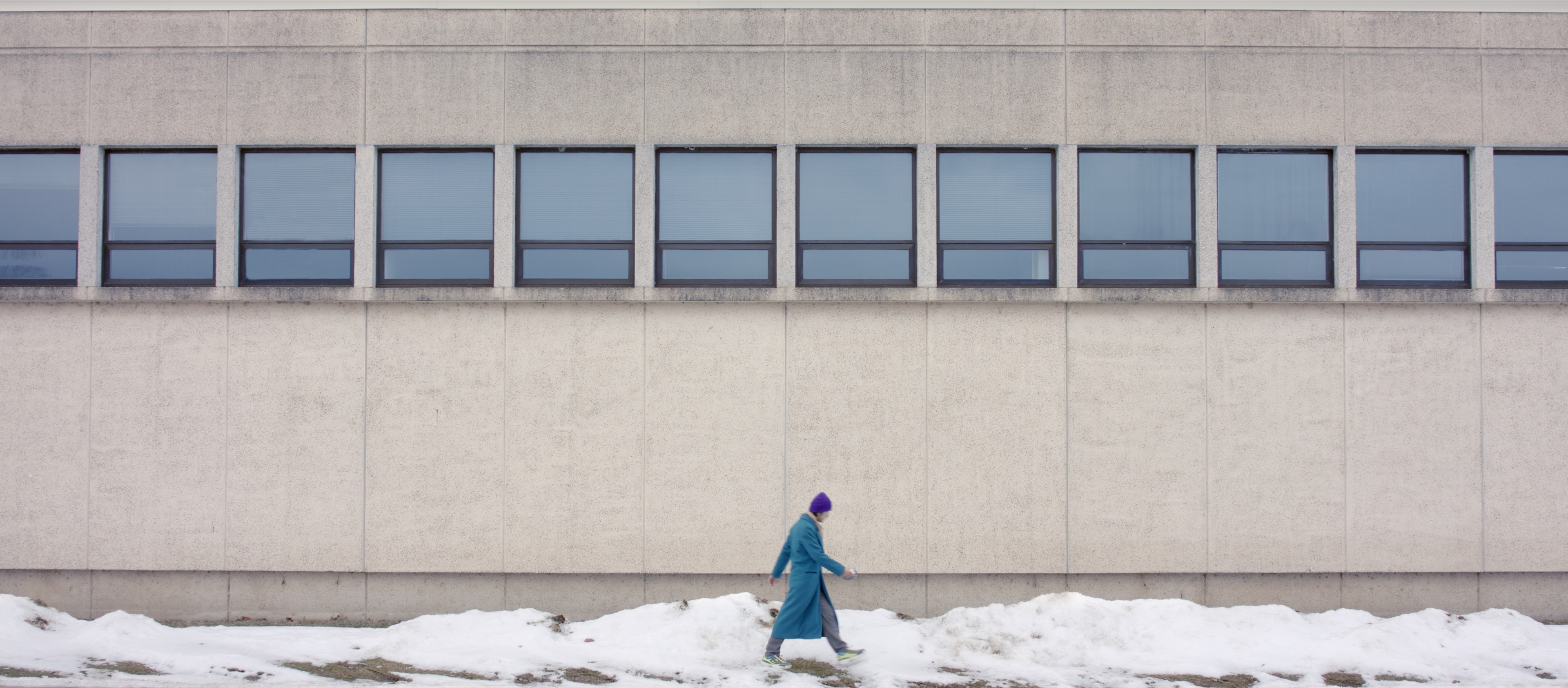 A solitary person in a blue coat and purple hat walking through snow beside a large concrete building.