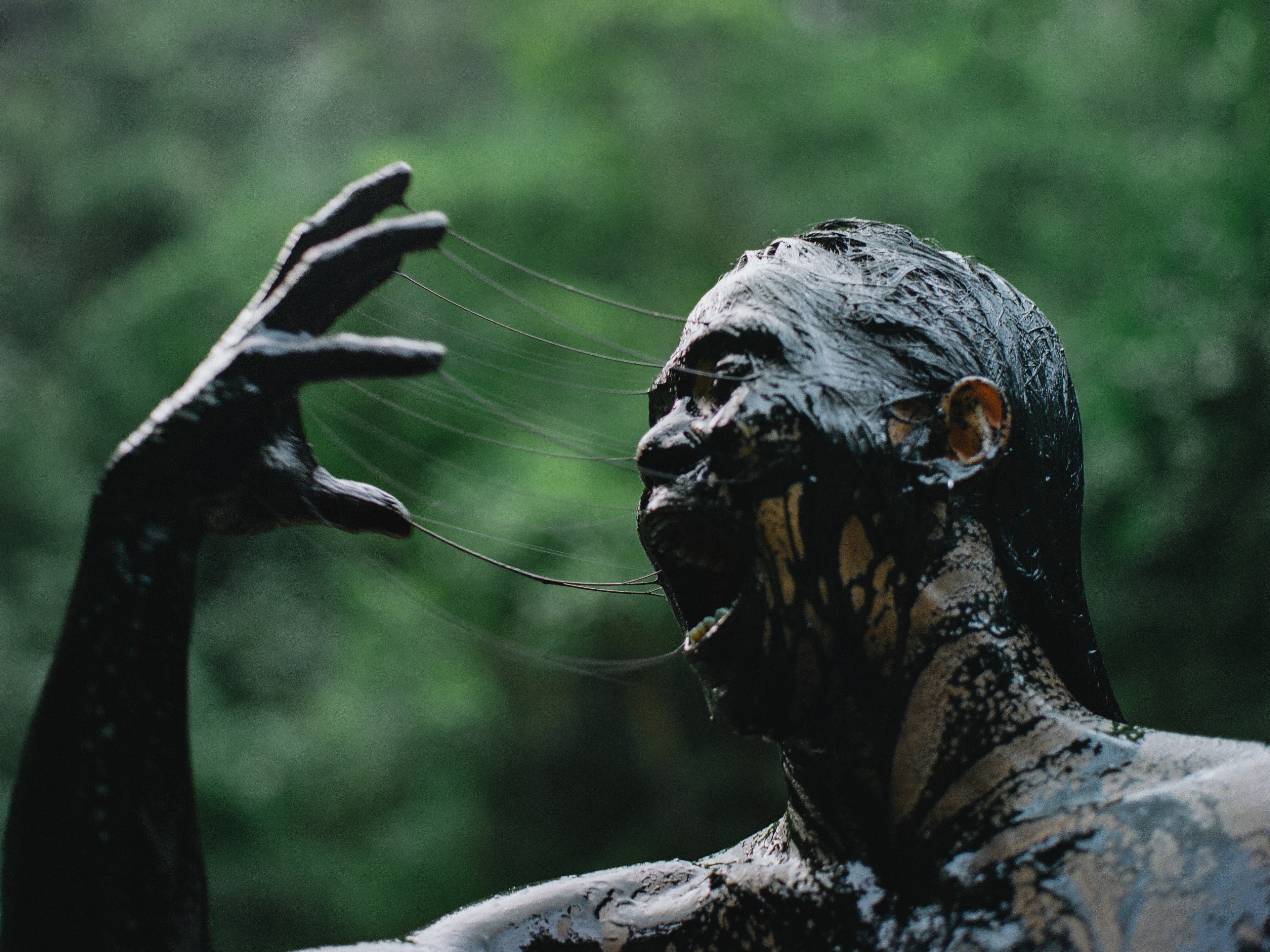 A figure covered in dark mud or paint stretching strands of sticky material between their hand and face against a blurred green background.