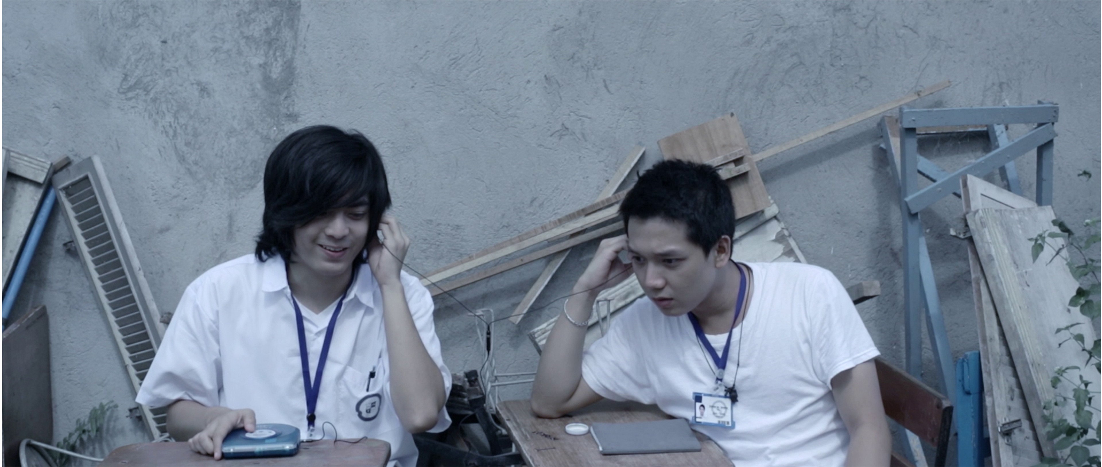 Two young men wearing ID lanyards sitting together at an outdoor table beside stacked construction materials.