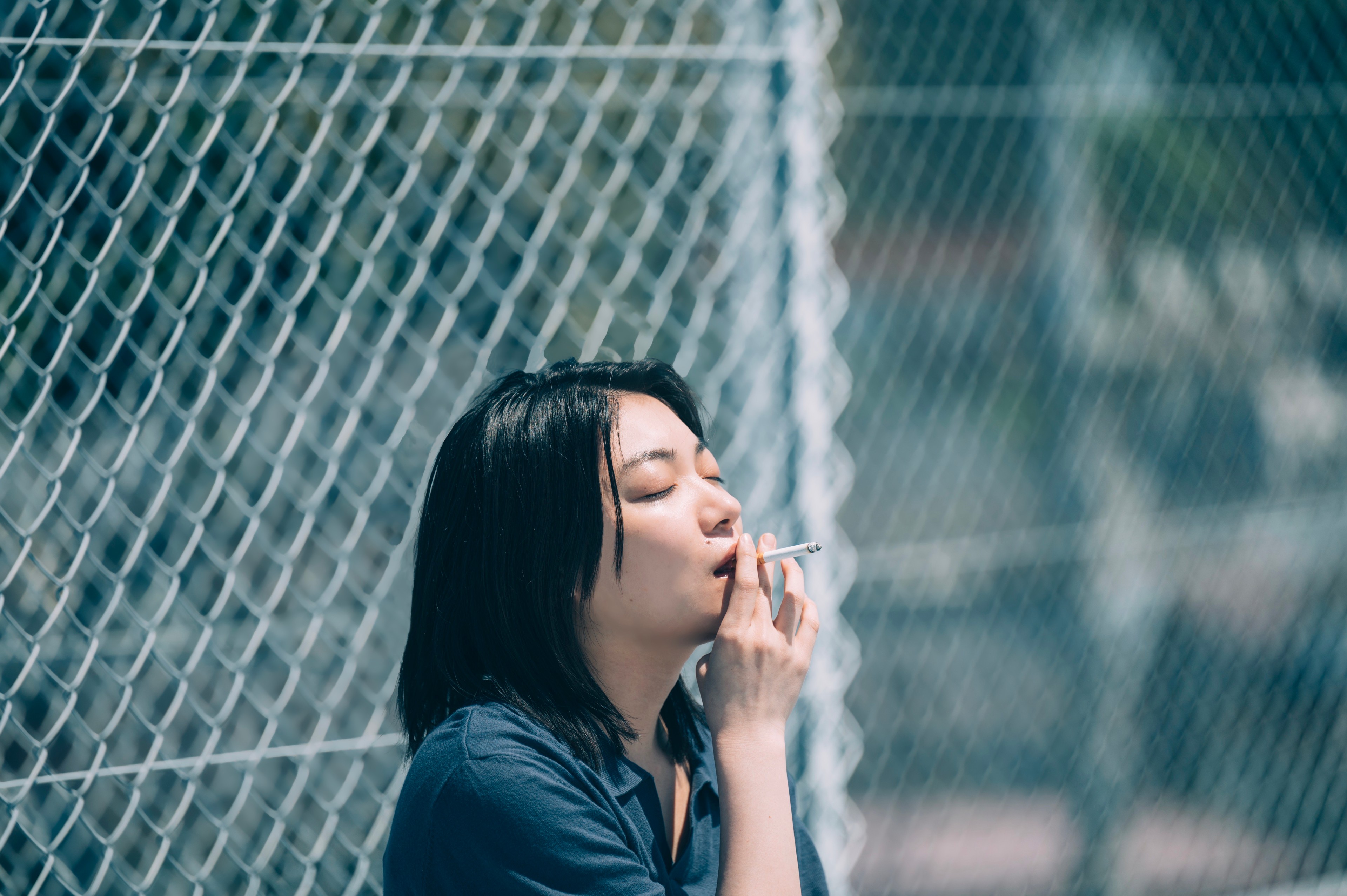 A woman smoking beside a chain-link fence in bright daylight.