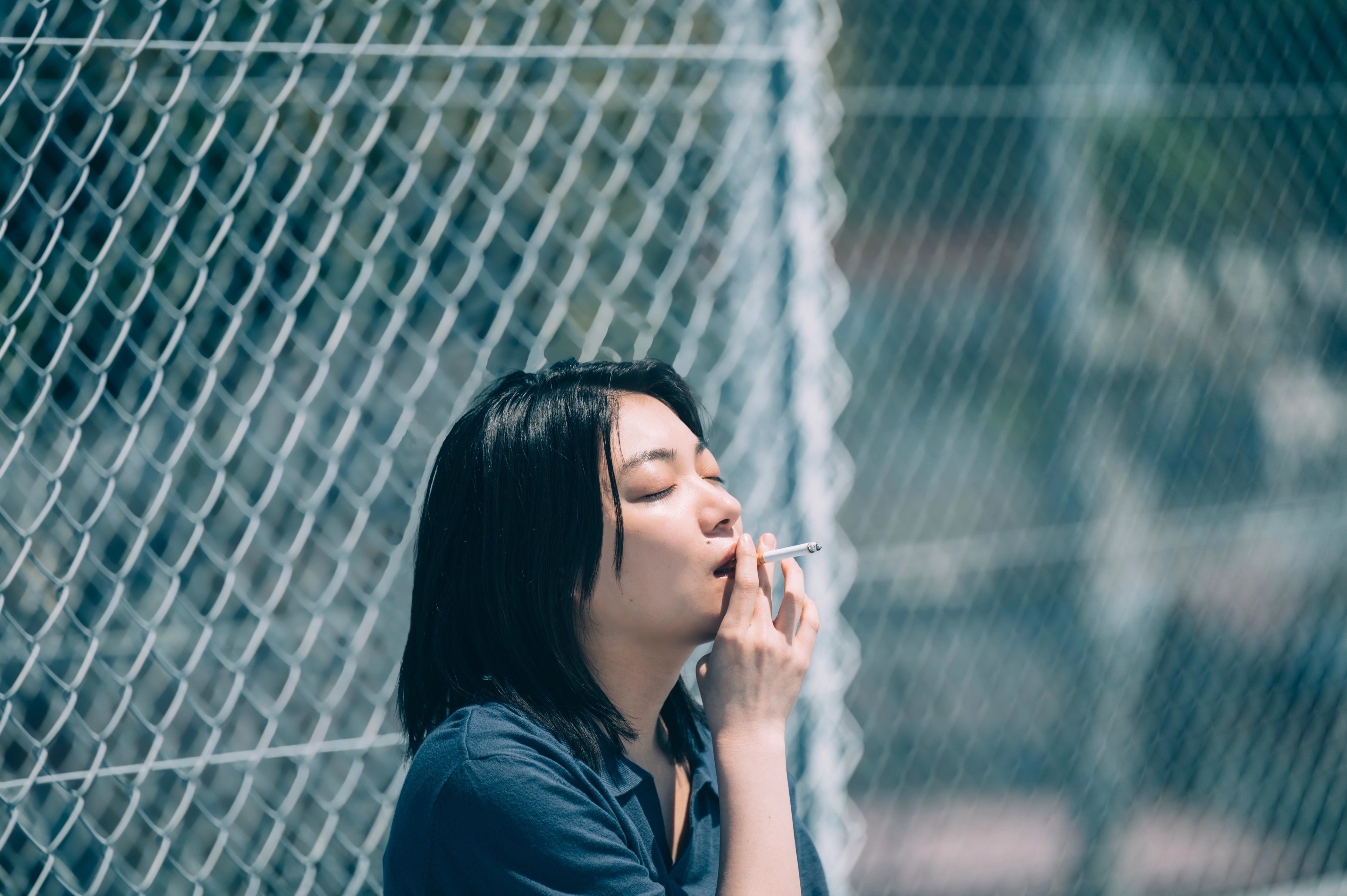 A woman smoking beside a chain-link fence in bright daylight.