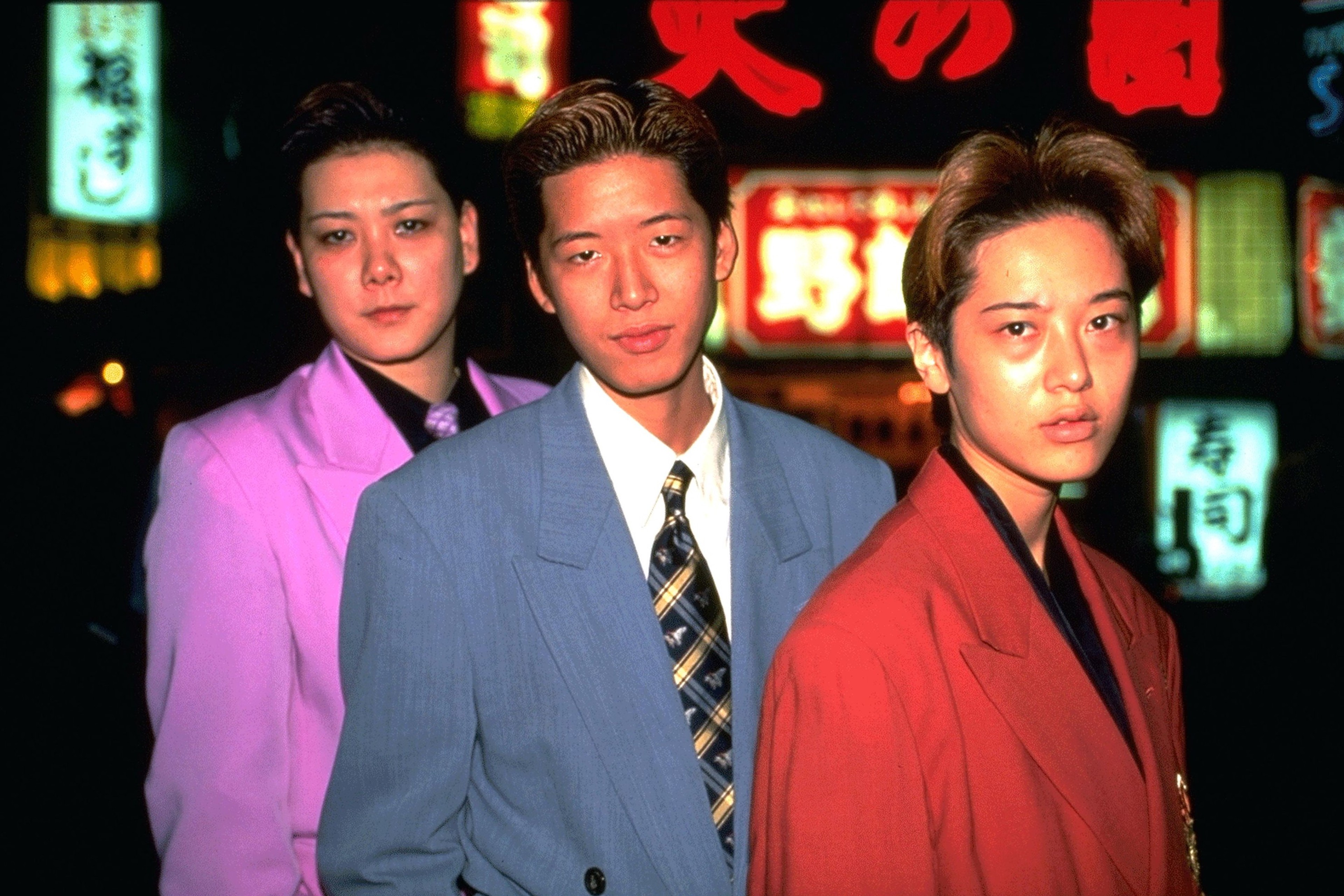 Three figures wearing suits stand against a backdrop of neon lights.