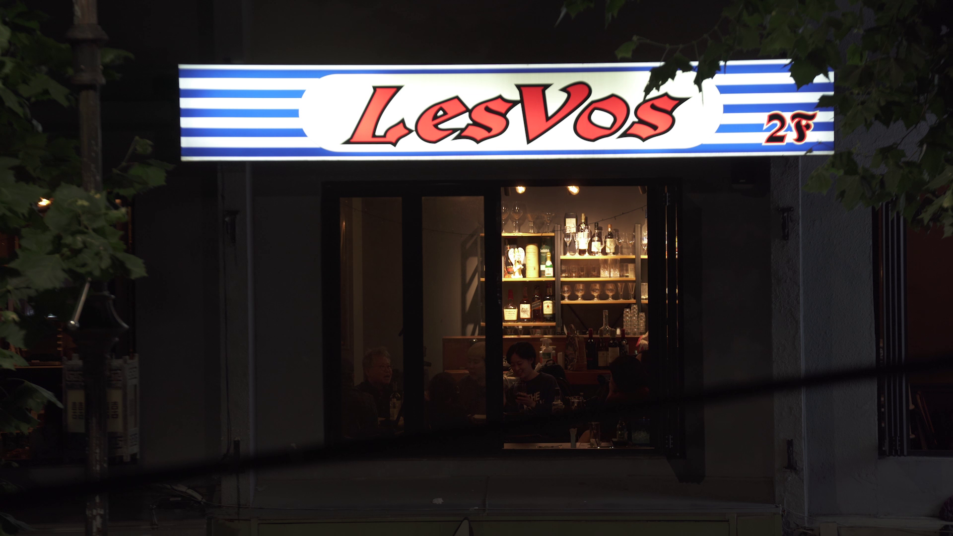 Exterior of a bar at night, with a brightly lit “LesVos” sign above the entrance and people visible inside.