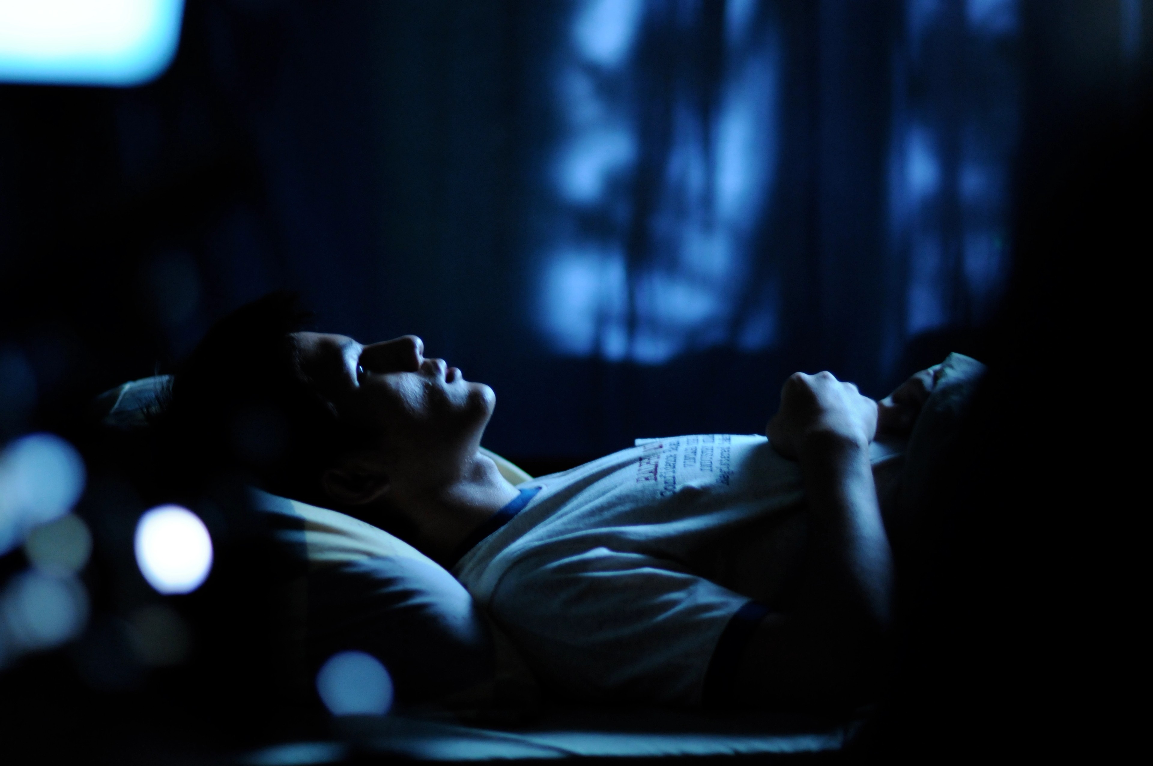A person lying on a bed in dim blue light, gazing upward in a shadowy room.