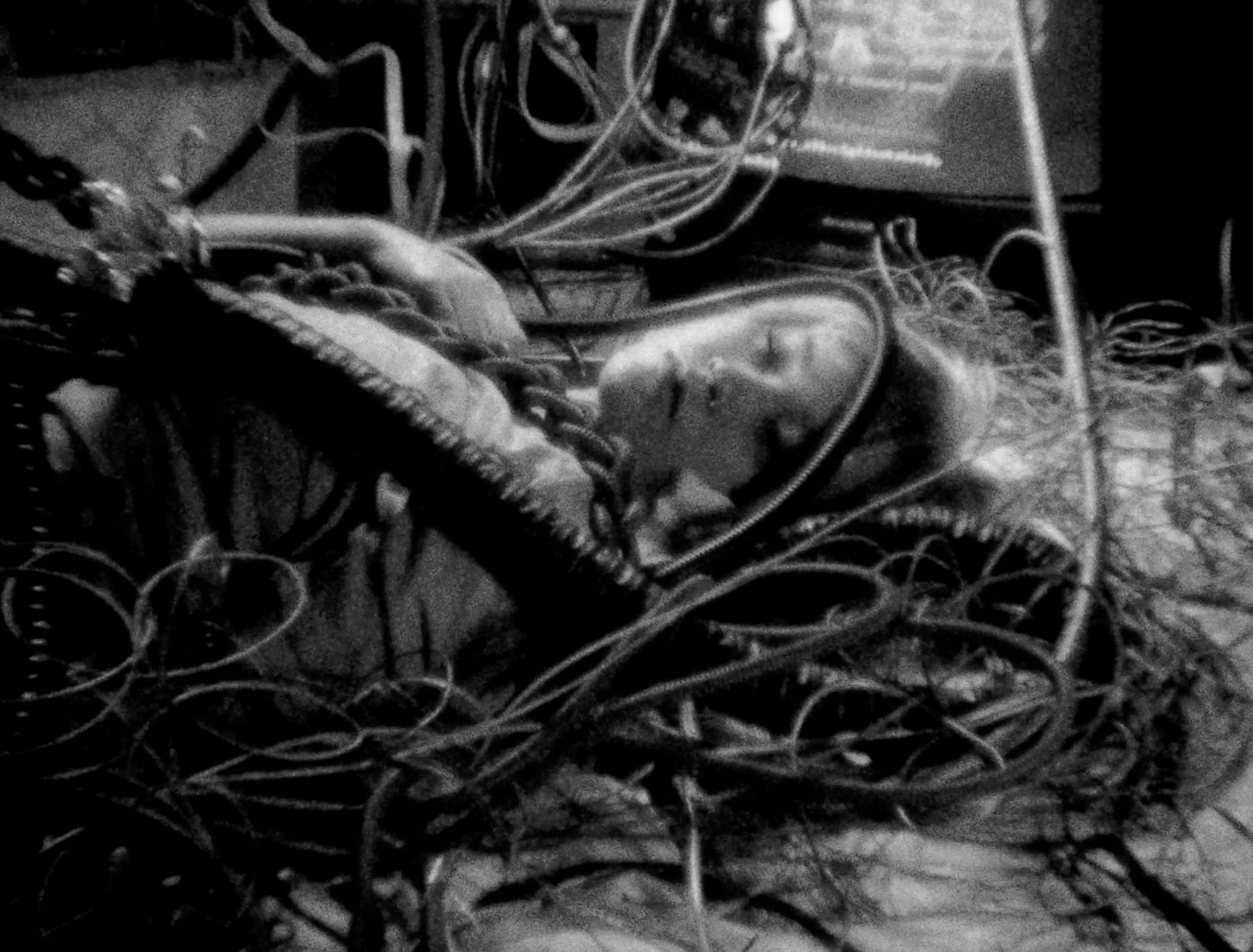 A person lying among tangled cables and electronic equipment in a grainy black-and-white image.