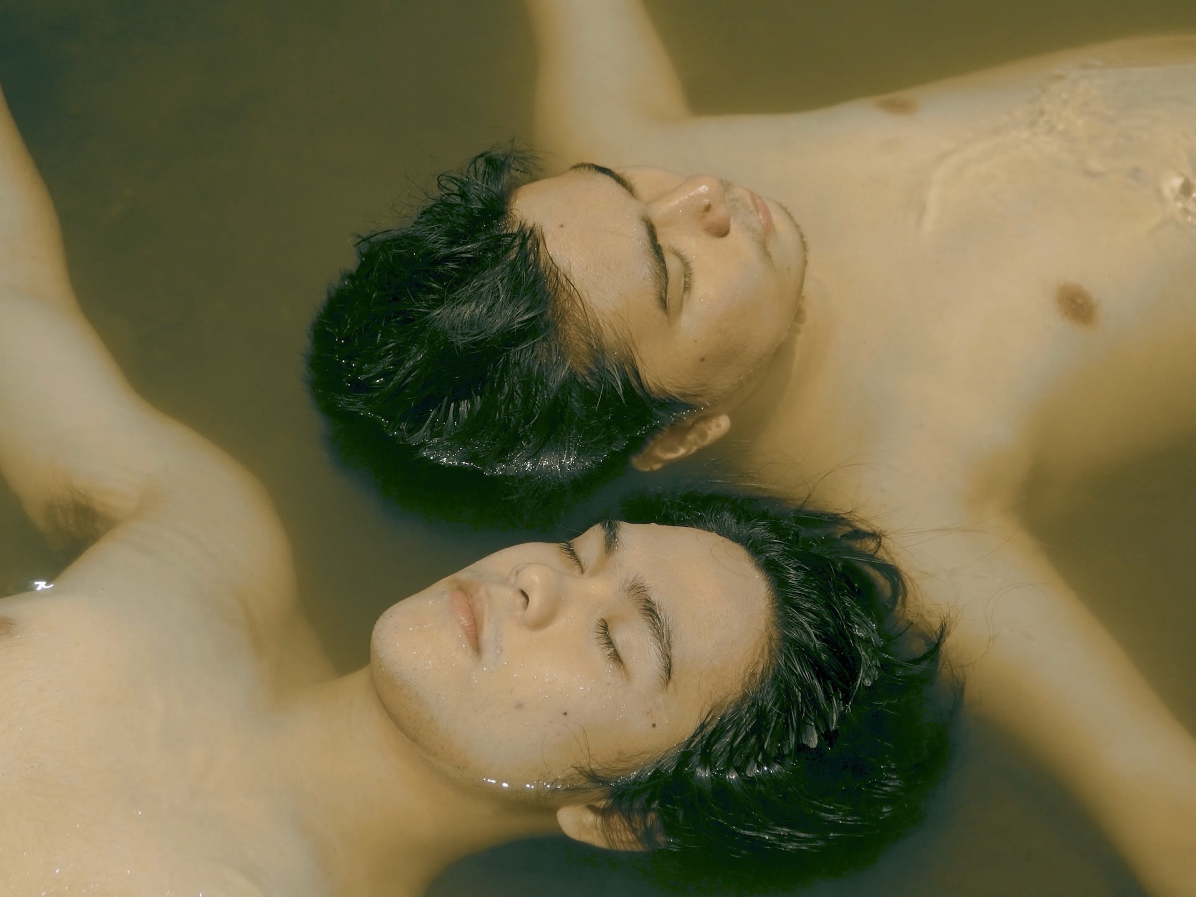 Two shirtless young men floating side by side in water with their eyes closed.