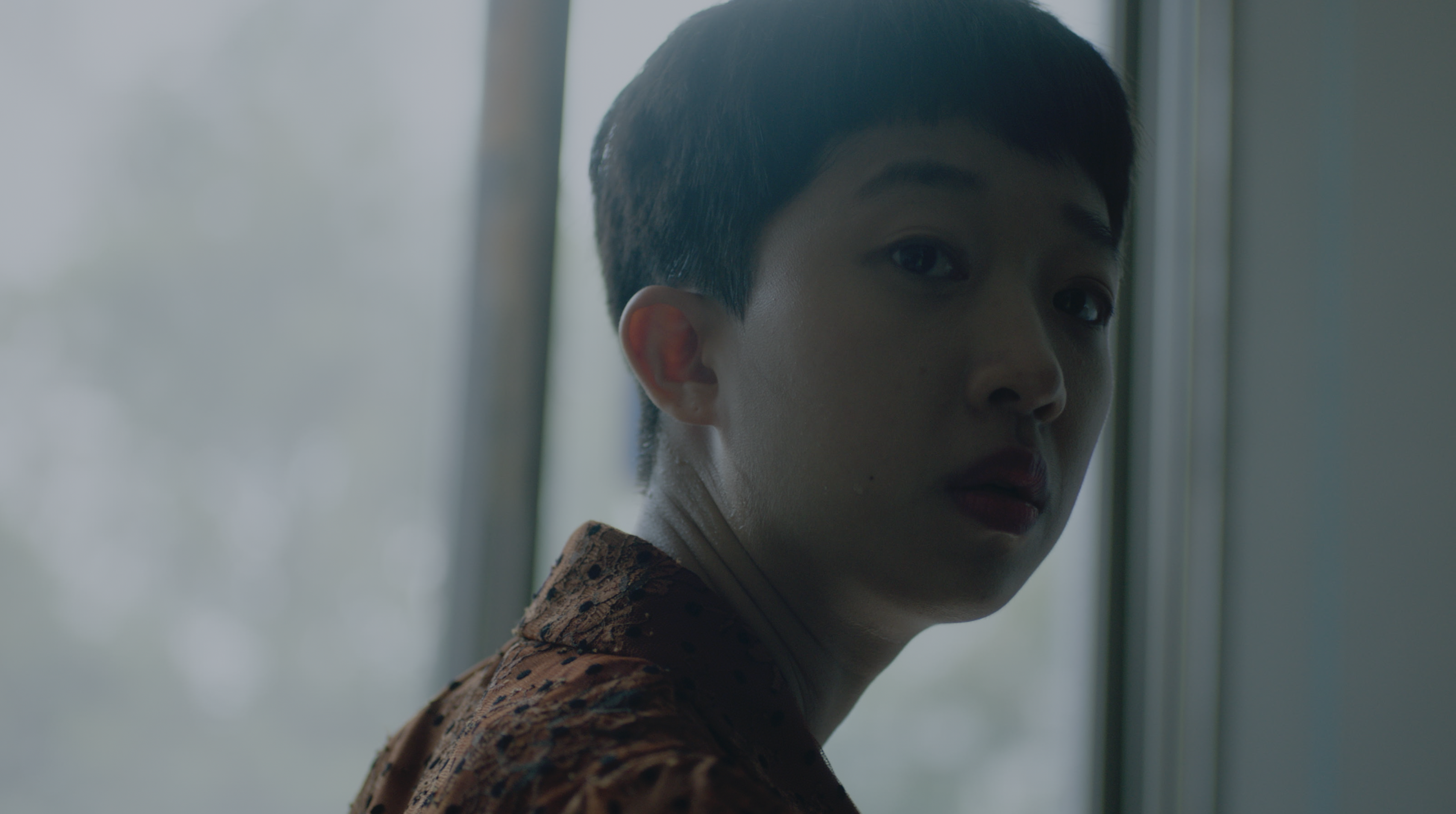 A young person with short hair looking back over their shoulder beside a softly lit window.