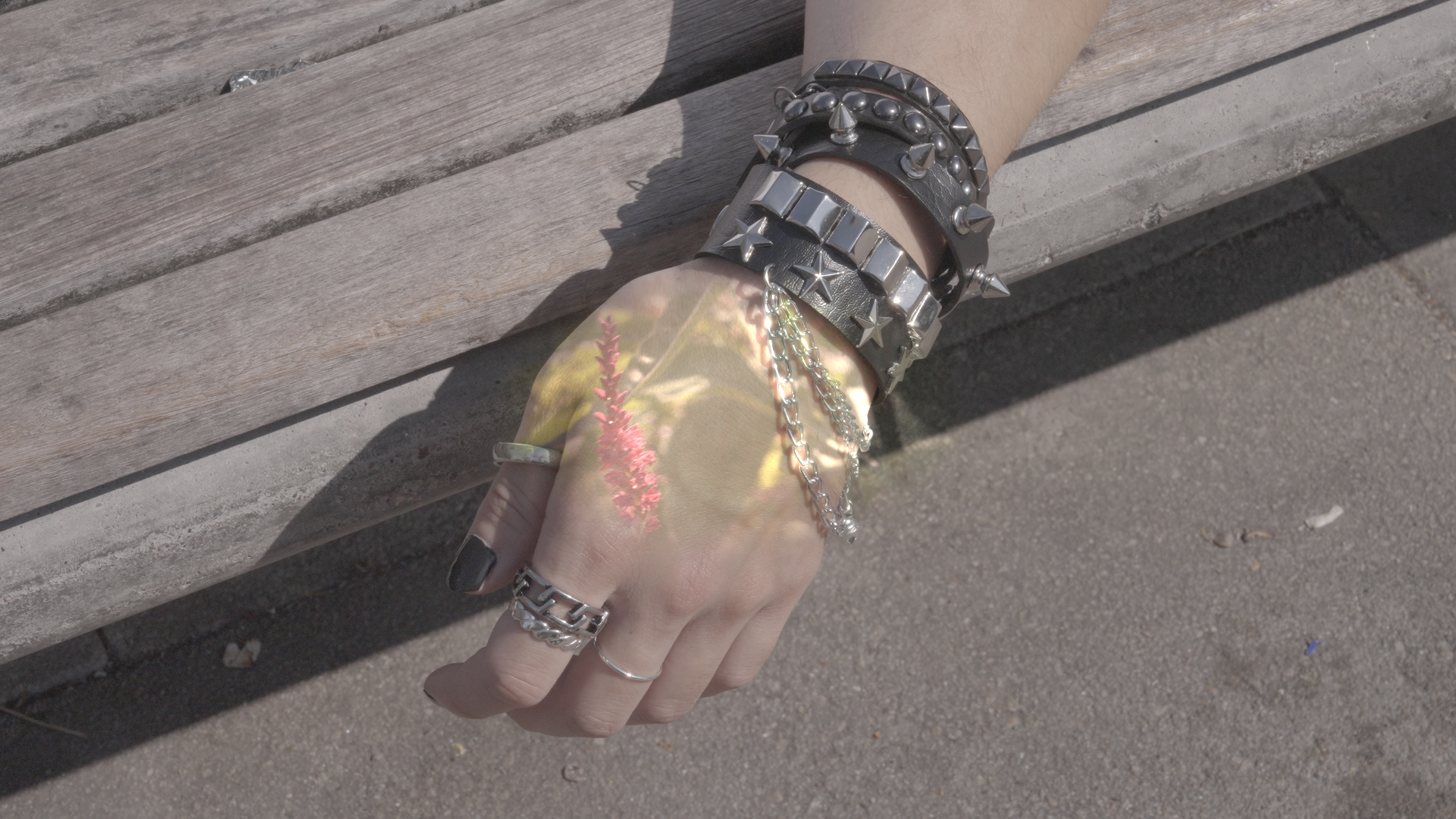 A hand adorned with rings, chains and studded bracelets rests beside a wooden bench, with delicate floral imagery projected across the skin.