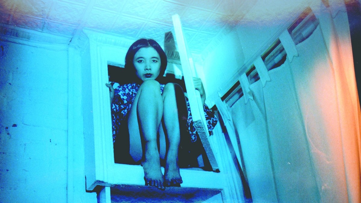 A young woman crouching on a narrow ledge in a small room illuminated by vivid blue light.